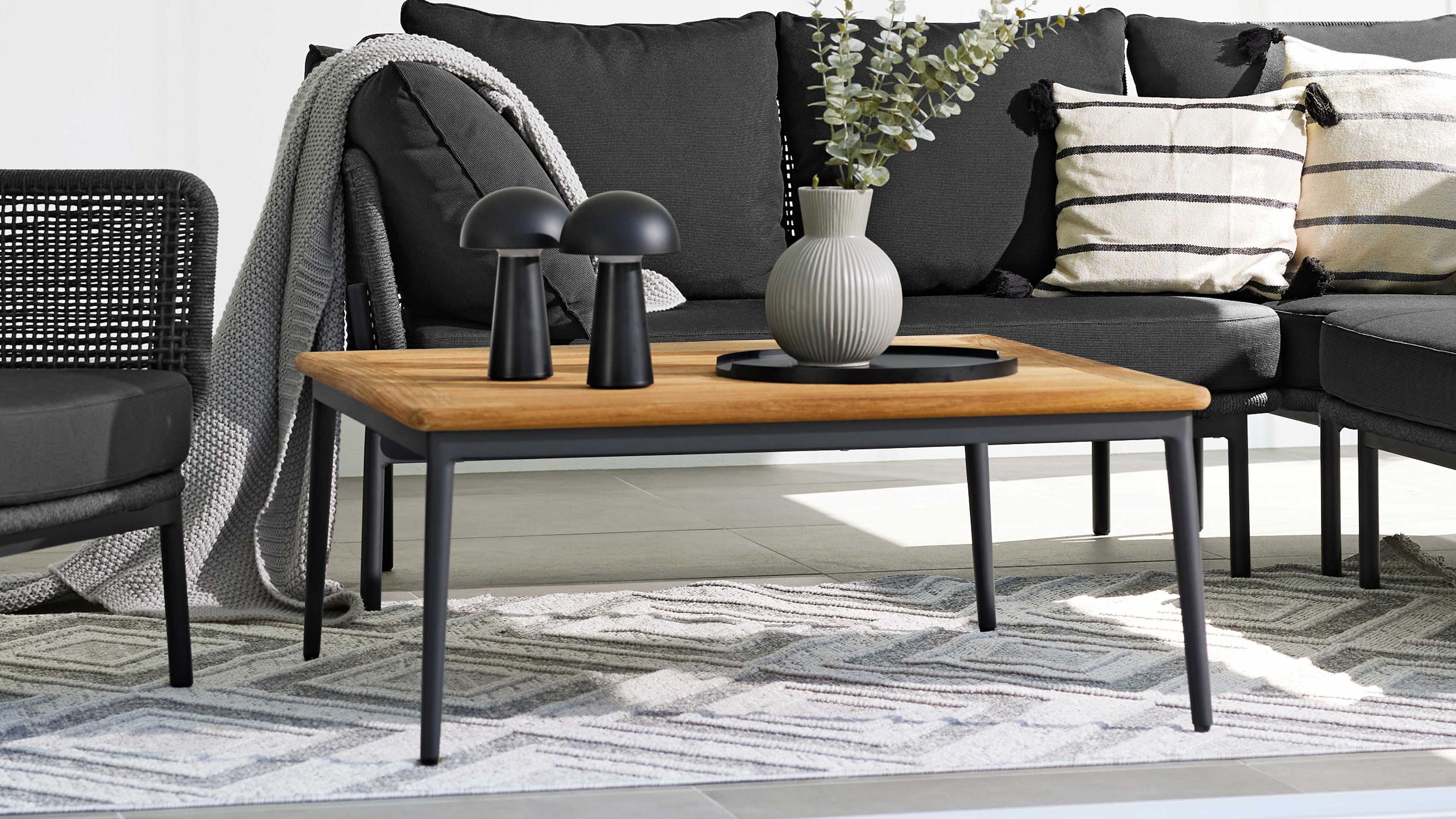 Quadratischer Lounge-Tisch mit hellem Holzplatte und schwarzen Metallbeinen vor schwarzer Sofalandschaft