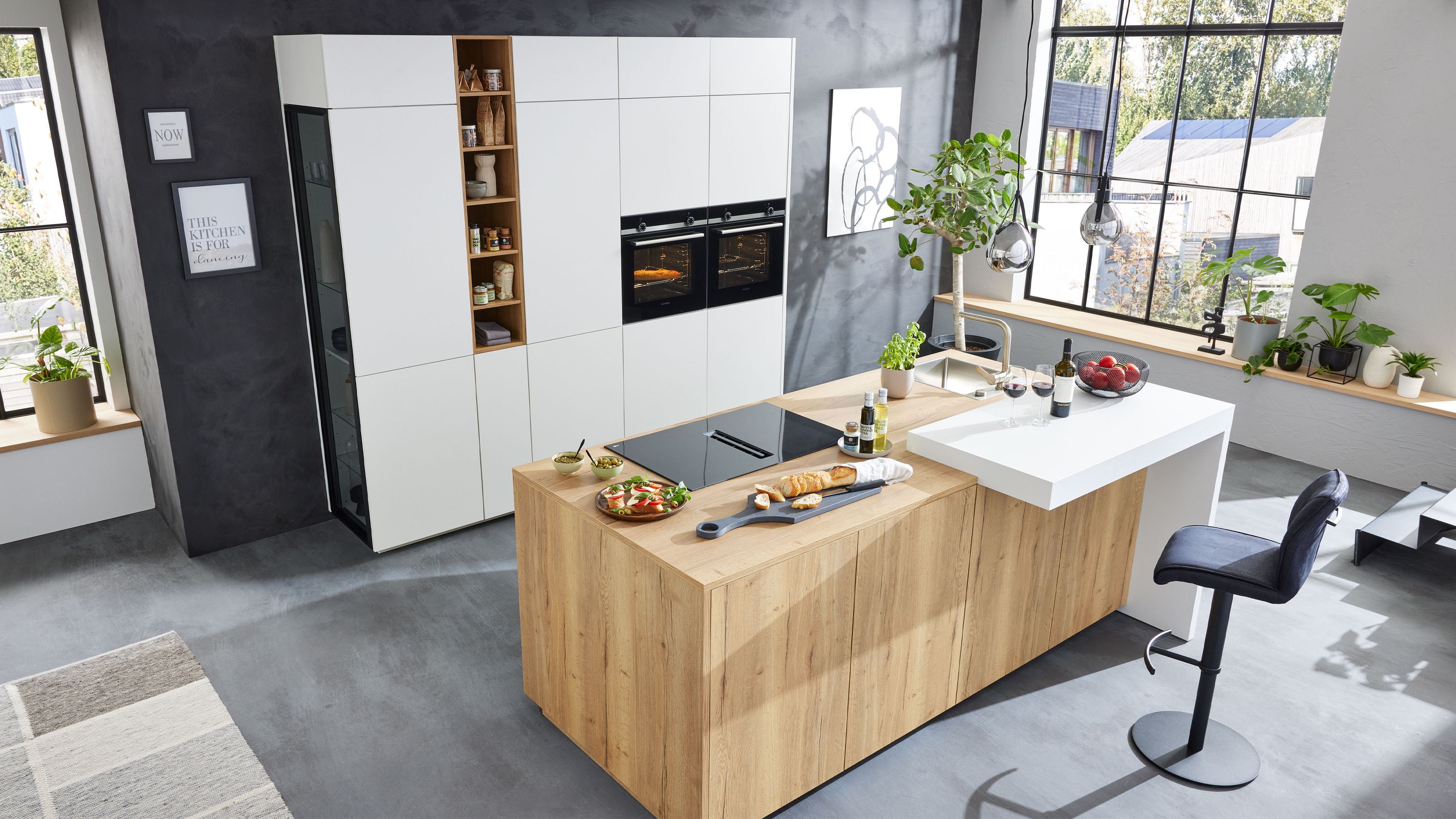 Moderne Küche mit Holzinsel integriertem Kochfeld weißen Hochschränken mit Einbaugeräten und großen Fenstern.