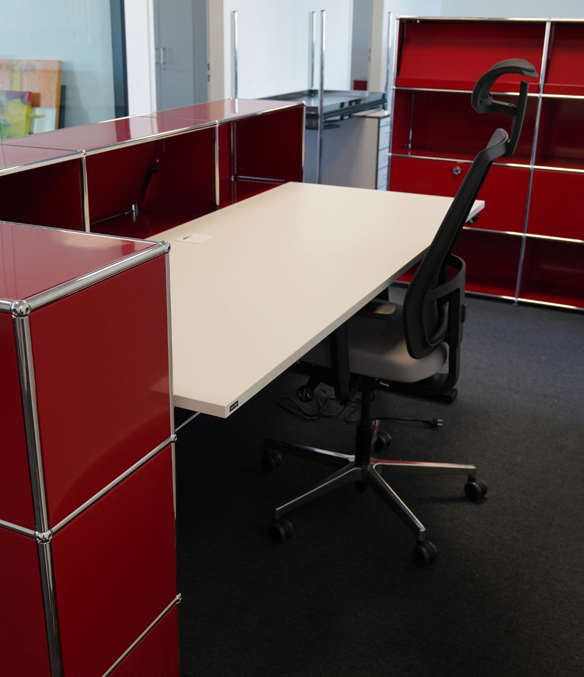 Moderner Arbeitsplatz mit Schreibtisch und Drehstuhl, umgeben von roten Trennwänden und Regalsystem im strukturierten Bürodesign