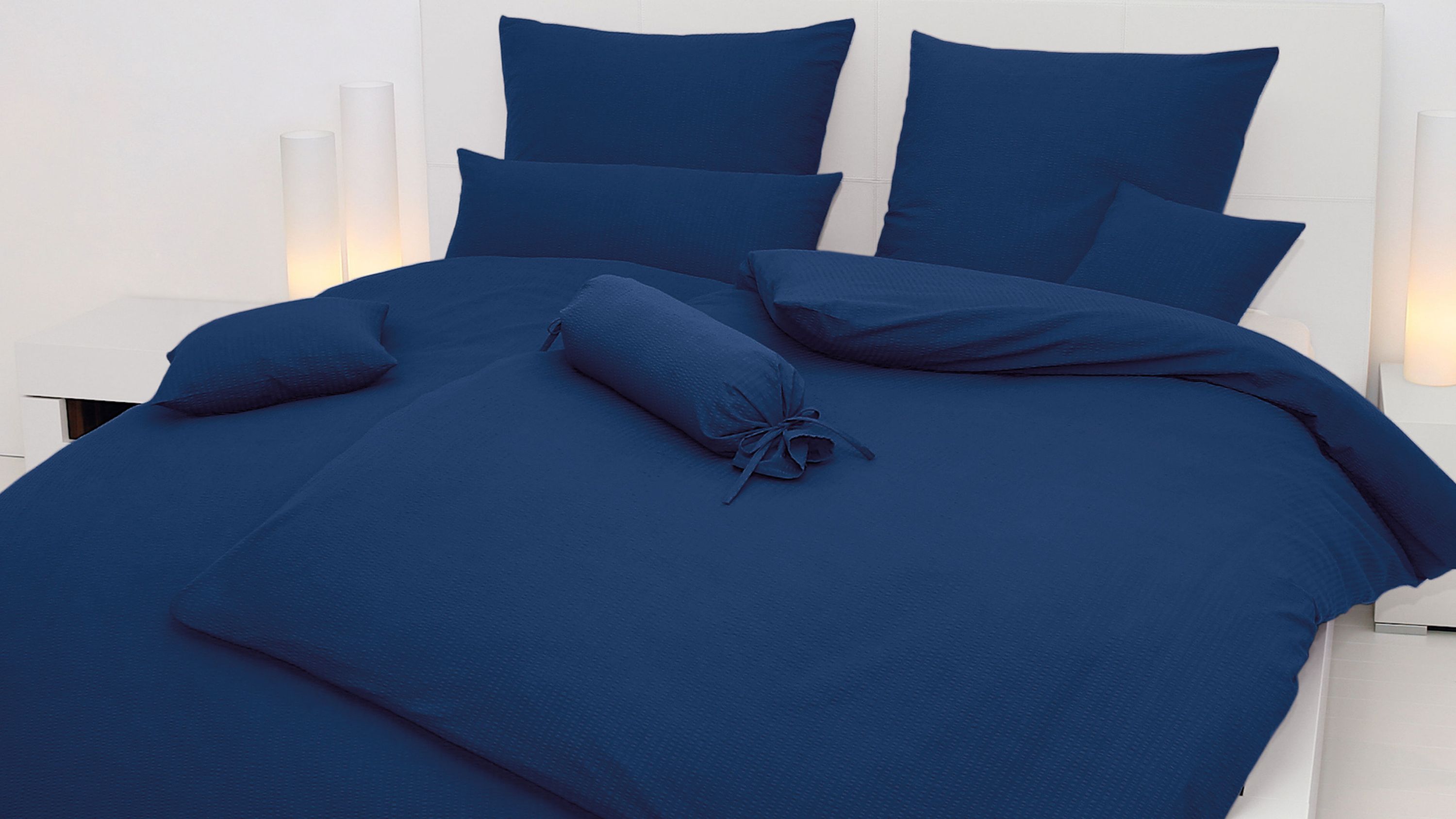 Dunkelblaue Bettwäsche mit Kissen und Zierrolle auf weißem Bett mit Kopfteil
