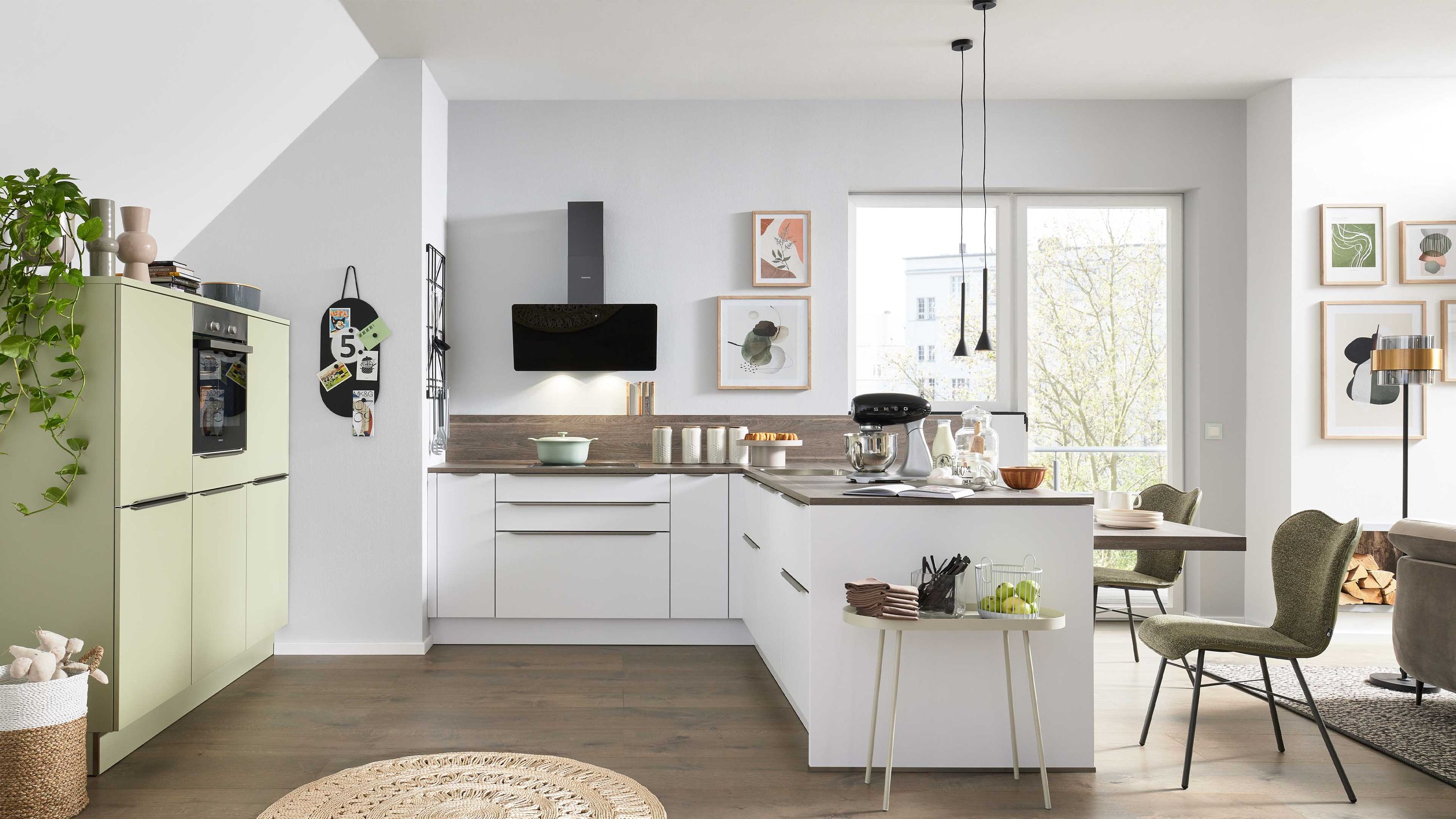 Moderne L-Küche in Weiß mit grünen Hochschränken Holzarbeitsplatte Einbauofen und großen Fenstern.