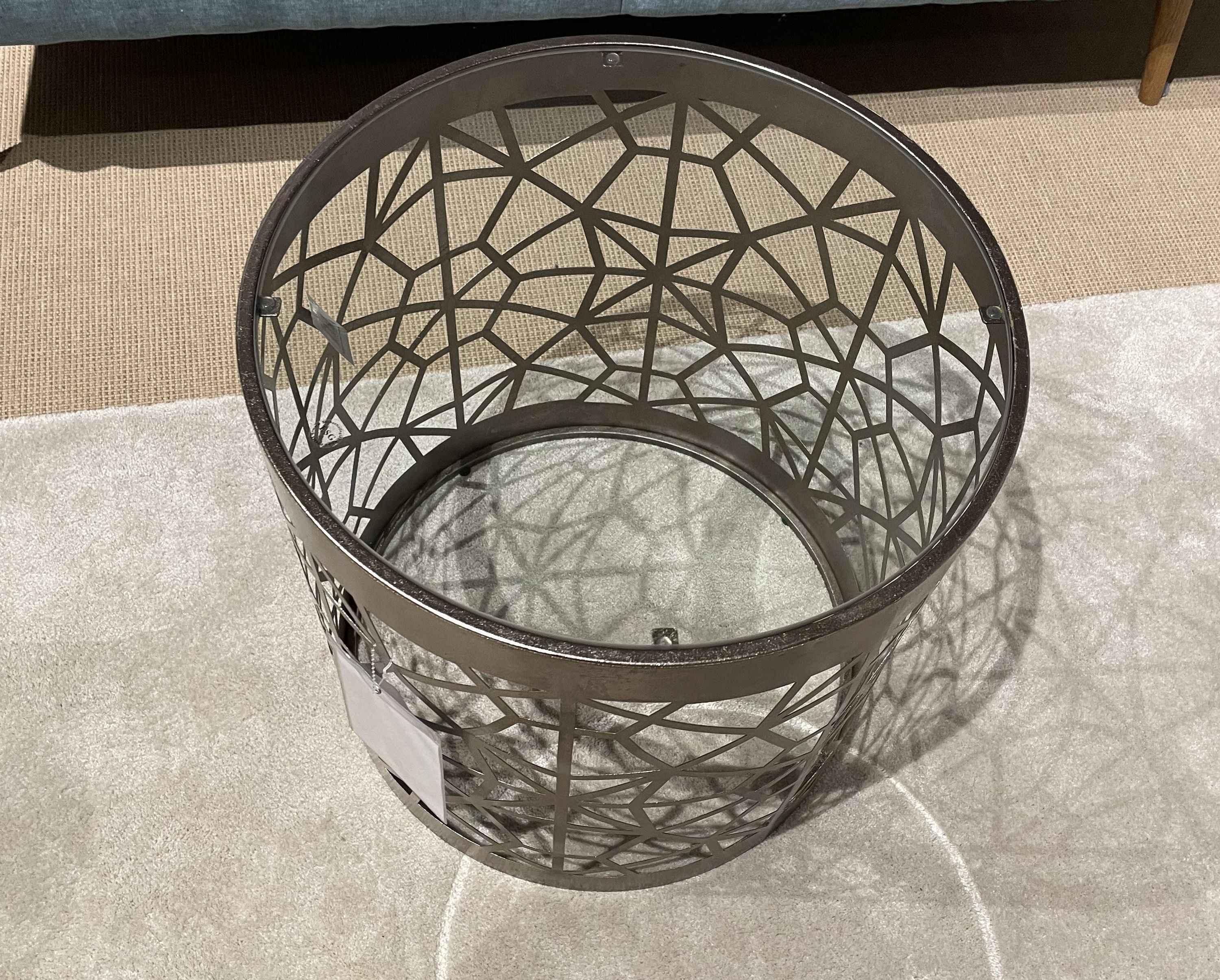 Runder Beistelltisch mit bronzefarbenem, geometrischem Metallgestell und Glasplatte auf hellgrauem Teppich.