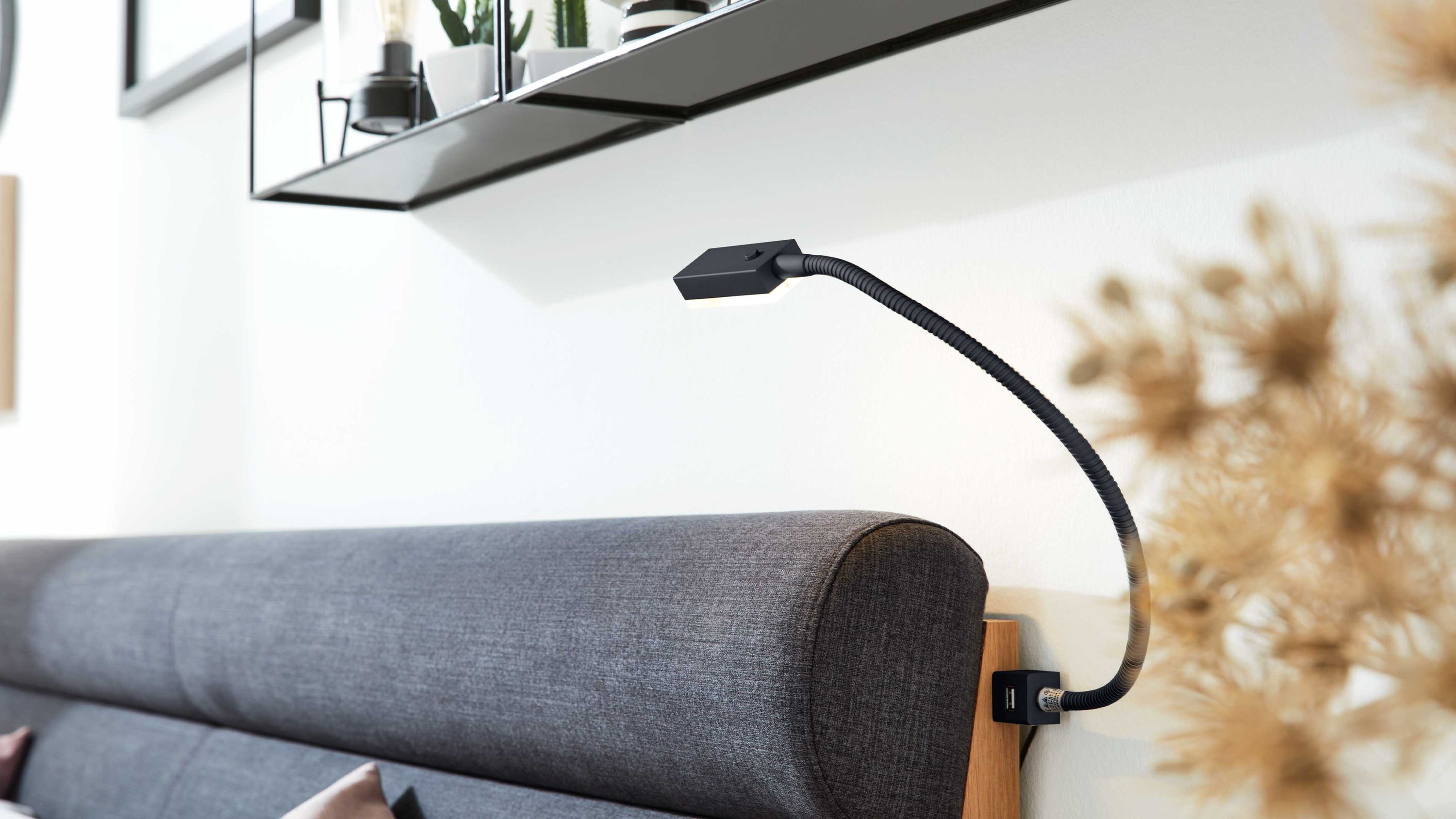 Schwarze flexible Leselampe mit kleinem rechteckigem Lampenkopf an einem grauen Polsterbett befestigt