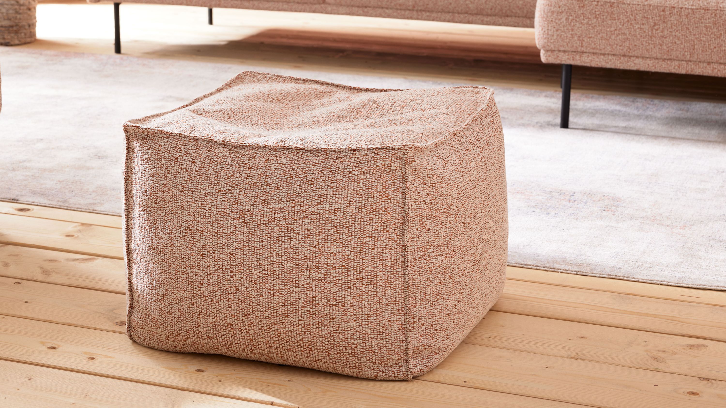Quadratischer Hocker mit rostfarben meliertem Stoffbezug auf Holzboden vor Sofa