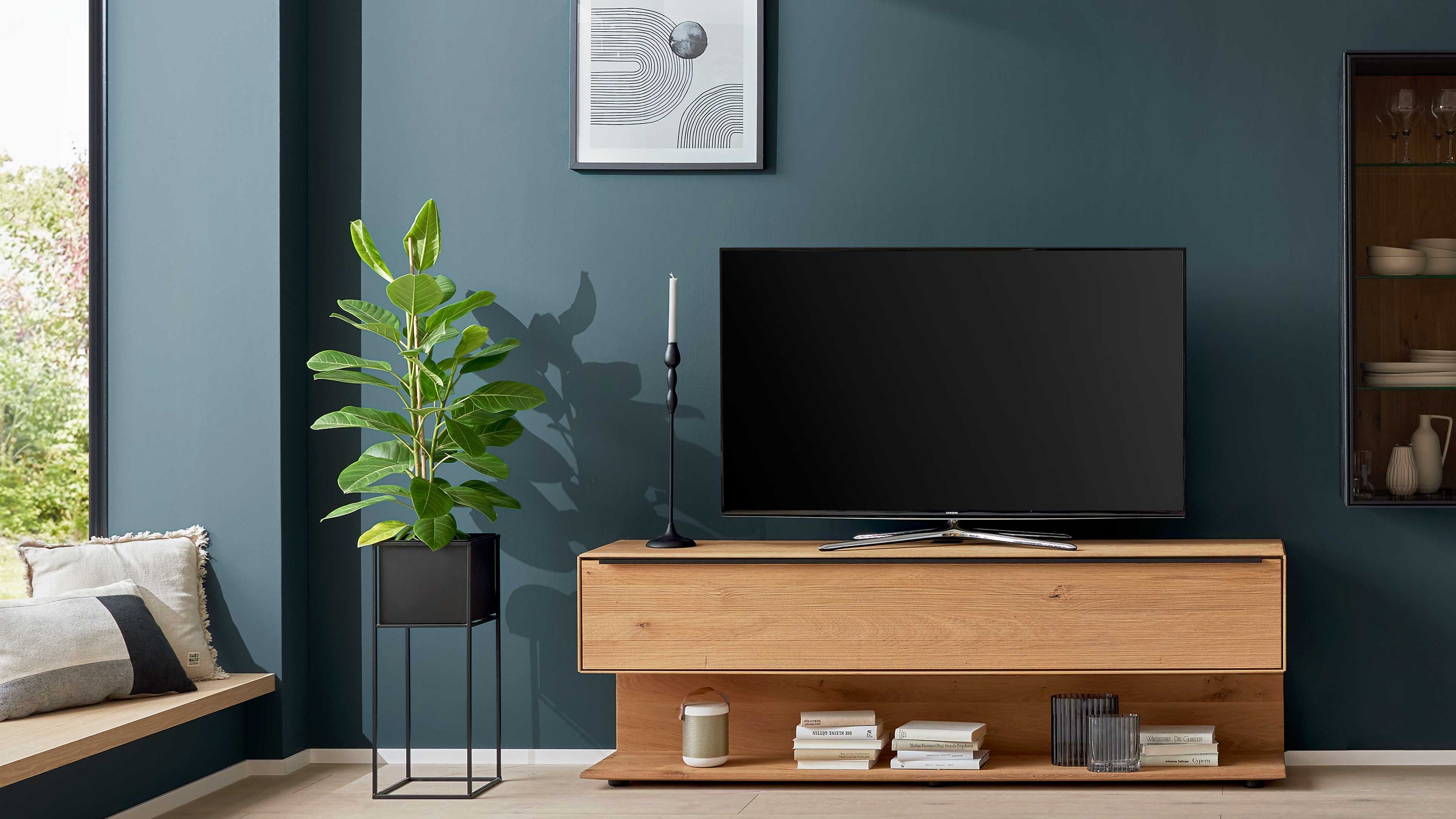 Lowboard mit Klapptür in Holzoptik Lowboard mit einer Klapptür in heller Holzoptik vor dunkelblauer Wand mit Fernseher und Pflanze