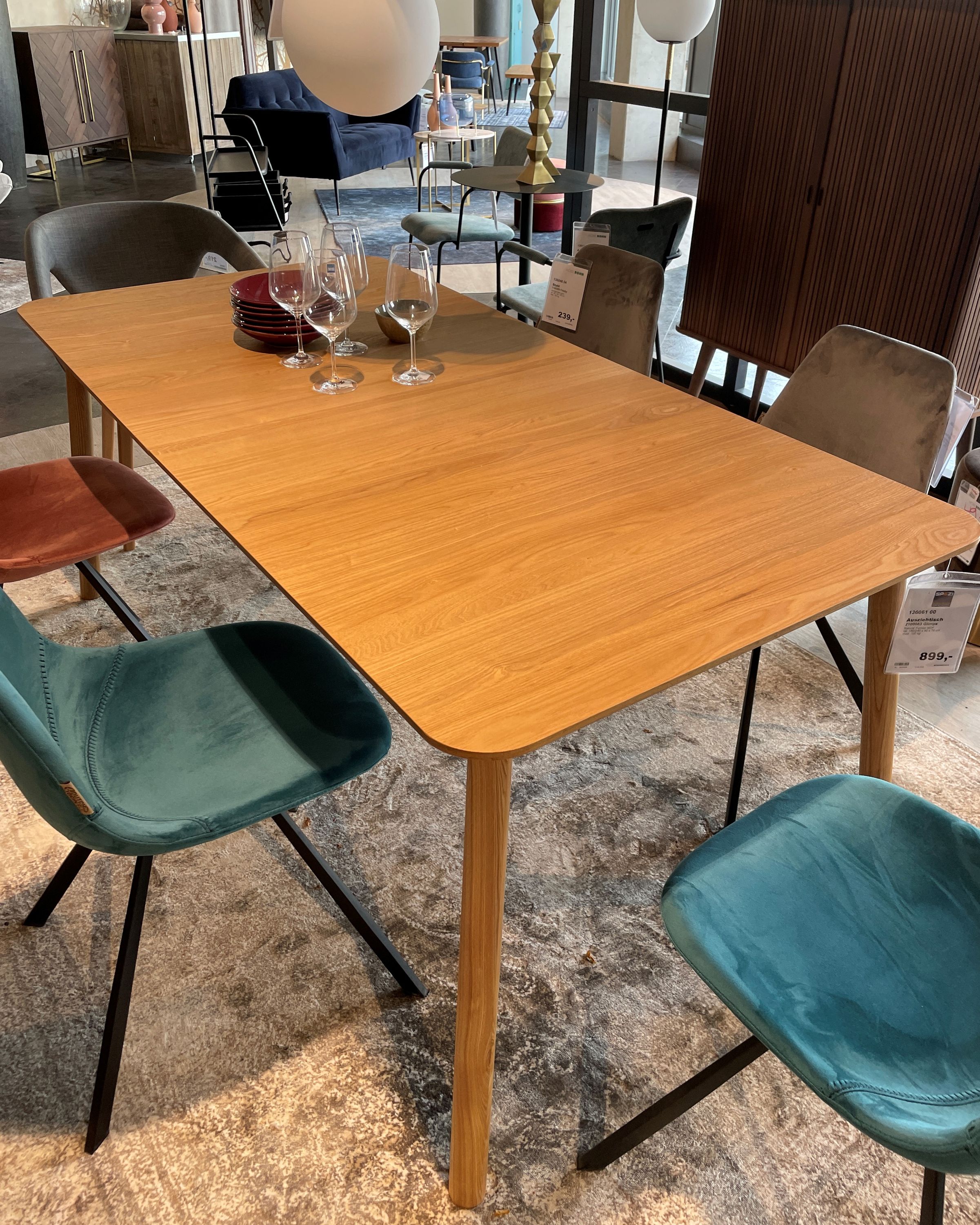 Rechteckiger Holztisch mit abgerundeten Ecken, umgeben von Samtstühlen in Blau, Grau und Rostrot, dekoriert mit Weingläsern.