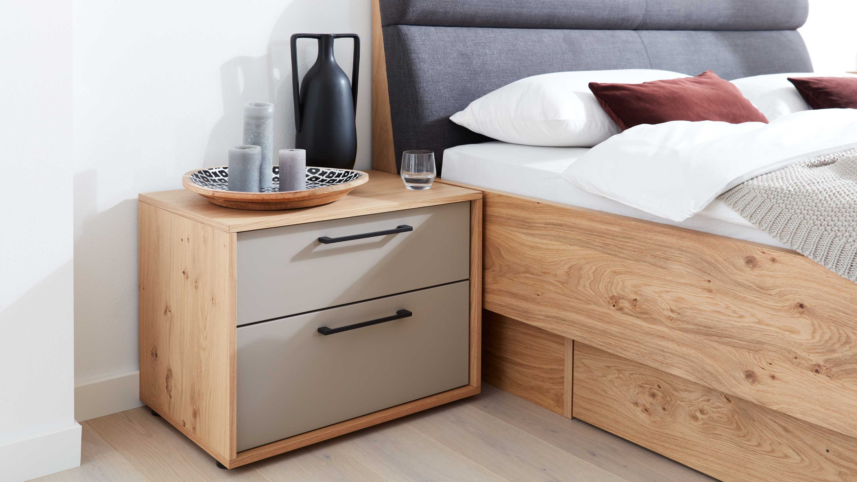 Nachtkommode mit zwei taupefarbenen Schubladen und Holzoptik neben einem Bett mit grauem Kopfteil