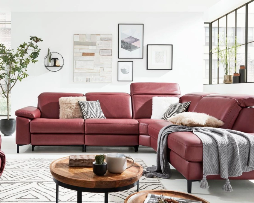 Gemütliches Wohnzimmer mit rotem Polstersofa, Dekokissen und stilvoller Wandgestaltung in warmem Ambiente. Wohnzimmer mit rotem Ecksofa, Couchtisch aus Holz, Wandbildern, Teppich in Naturtönen und großer Zimmerpflanze