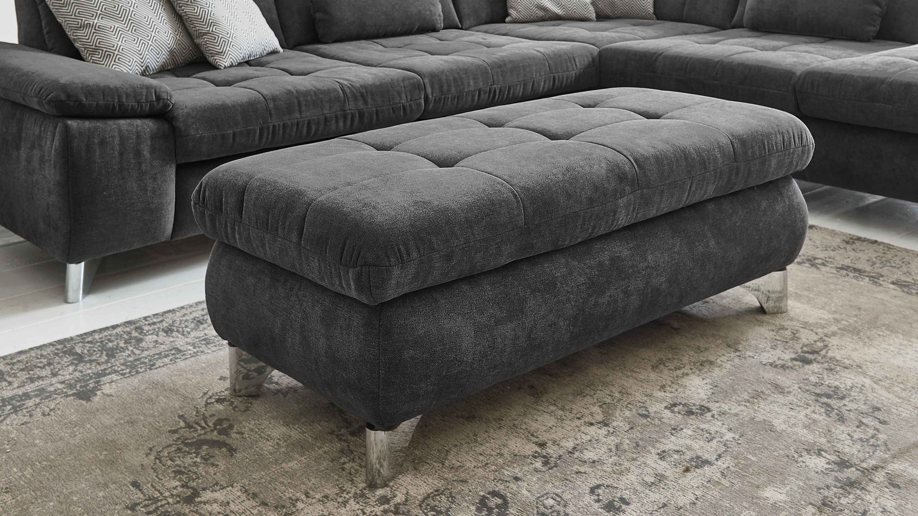 Großer rechteckiger Hocker mit schieferfarbenem Stoffbezug und glänzenden Metallfüßen auf Teppich vor dunklem Sofa