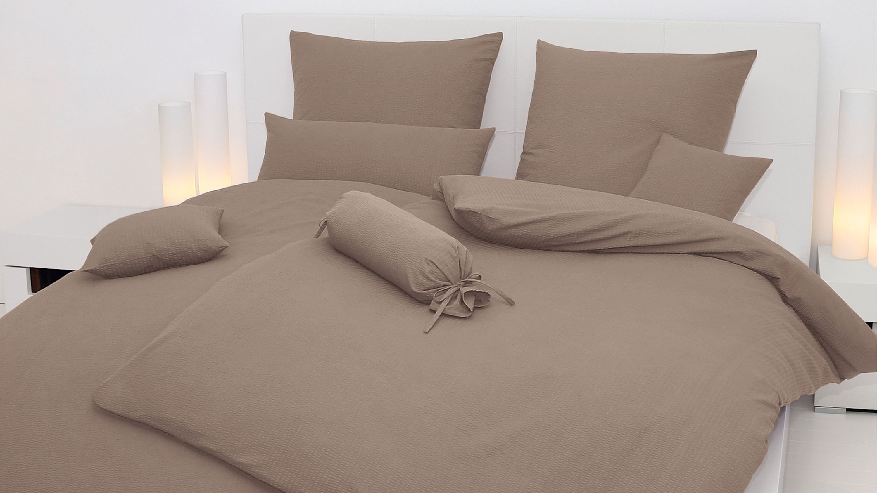 Taupefarbene Bettwäsche mit Kissen und Zierrolle auf weißem Bett in modernem Schlafzimmer