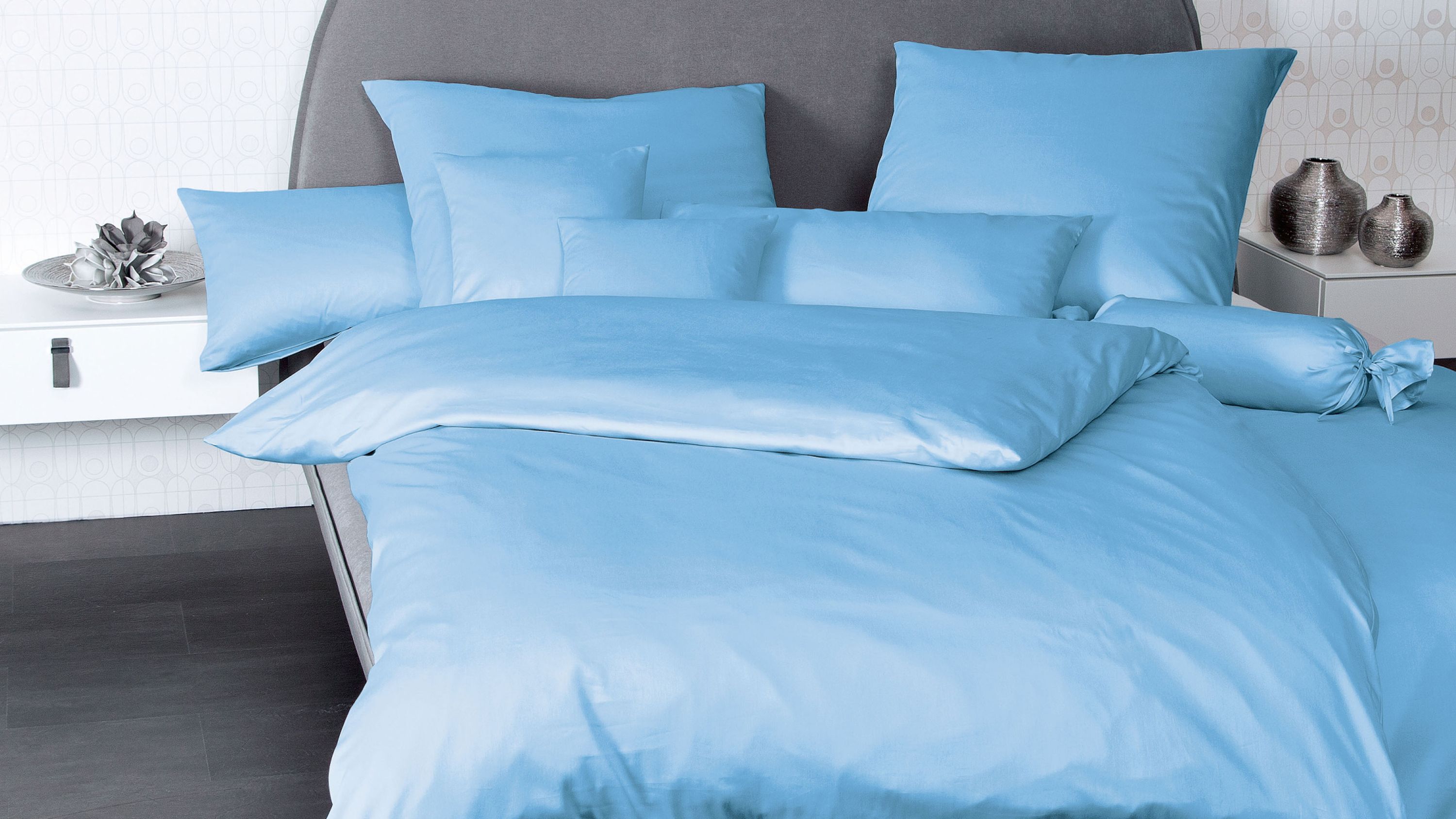 Hellblaue Bettwäsche mit mehreren Kissen und einer runden Nackenrolle auf einem grauen Bett