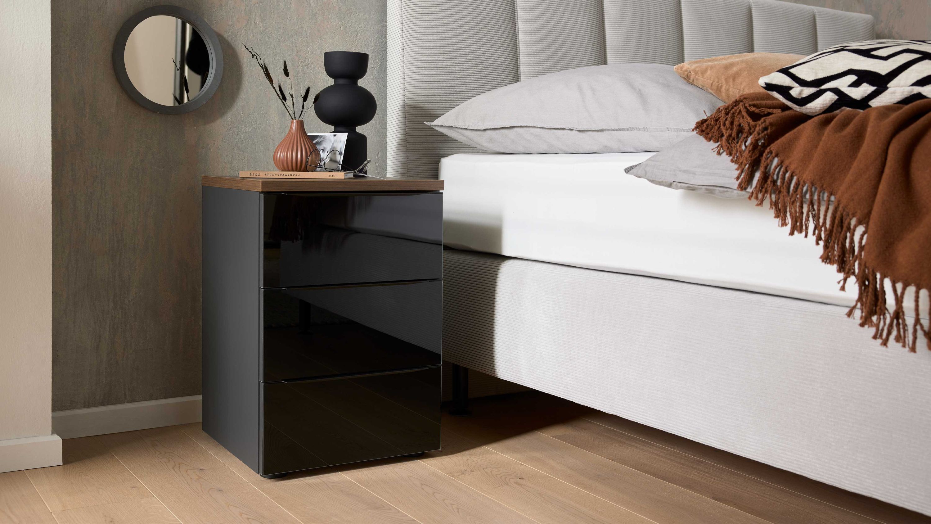 Schwarze Nachtkommode mit drei Schubladen und brauner Holzplatte neben einem grauen Bett mit Kissen und Decke
