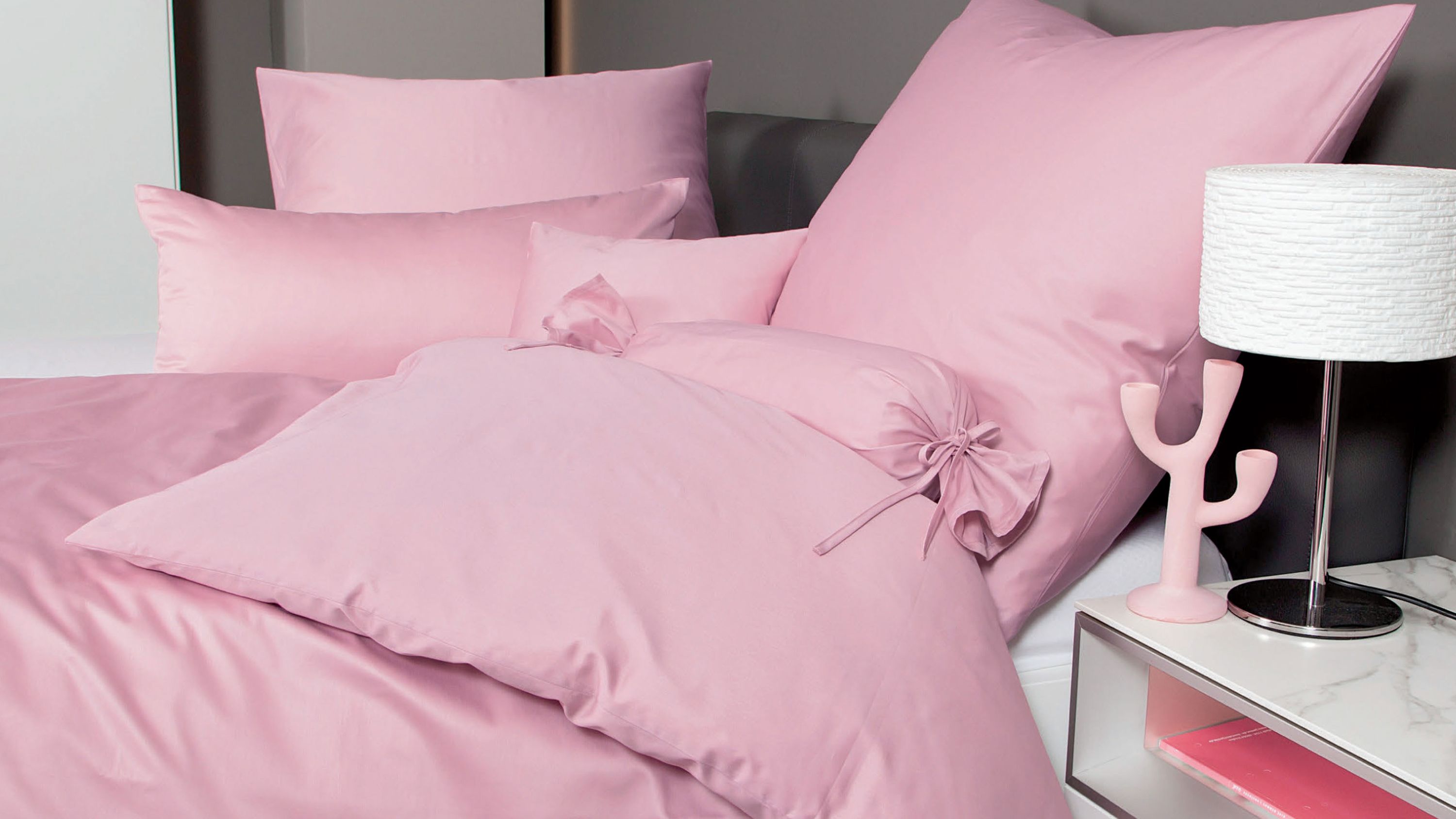 Rosa Bettwäsche-Set mit mehreren Kissen und Zierkissen auf einem Bett neben einem Nachttisch mit Lampe