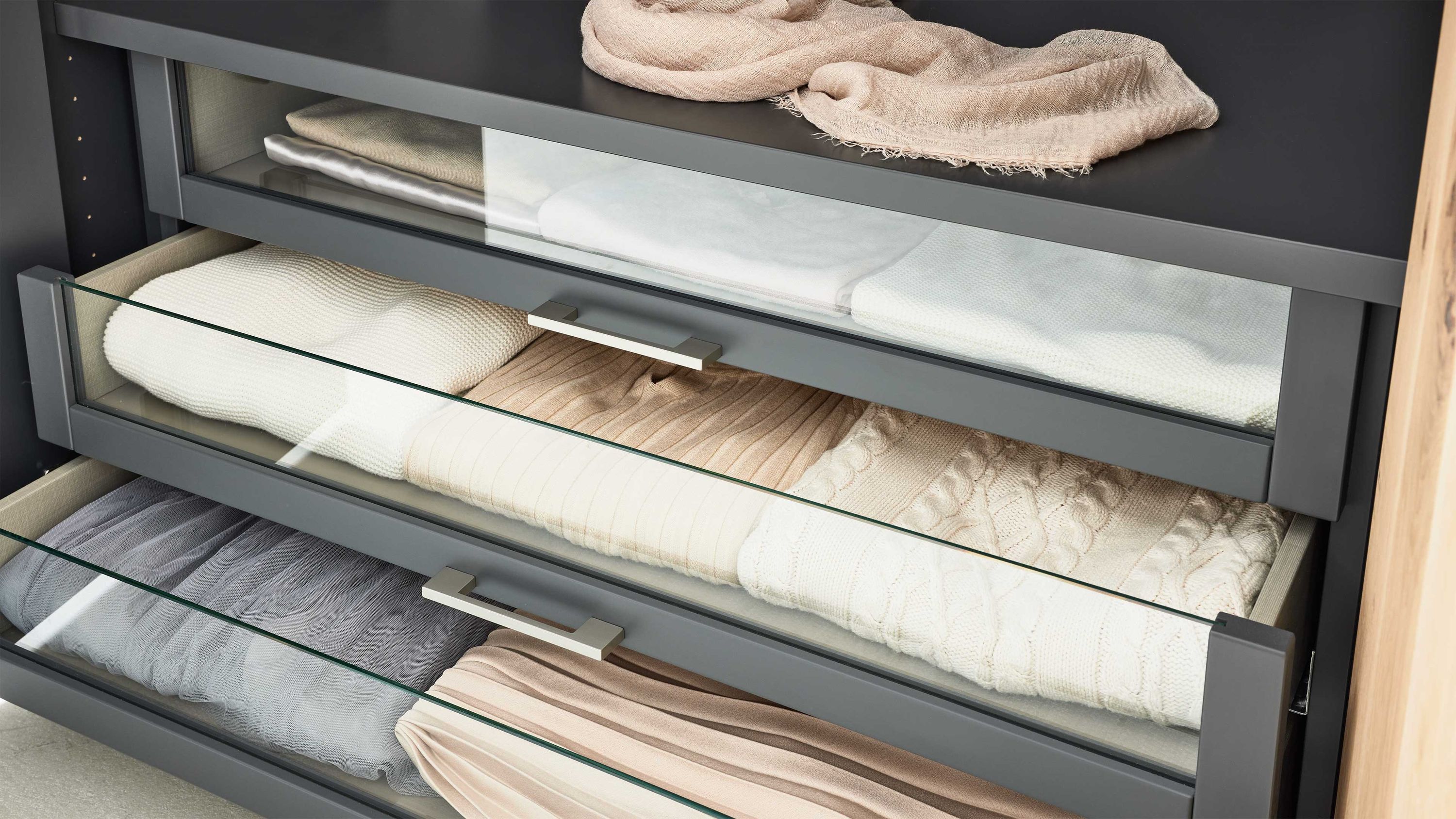 Drei graue Innenschubladen mit Glasfront gefüllt mit gefalteten Pullovern in Beige Weiß und Grau