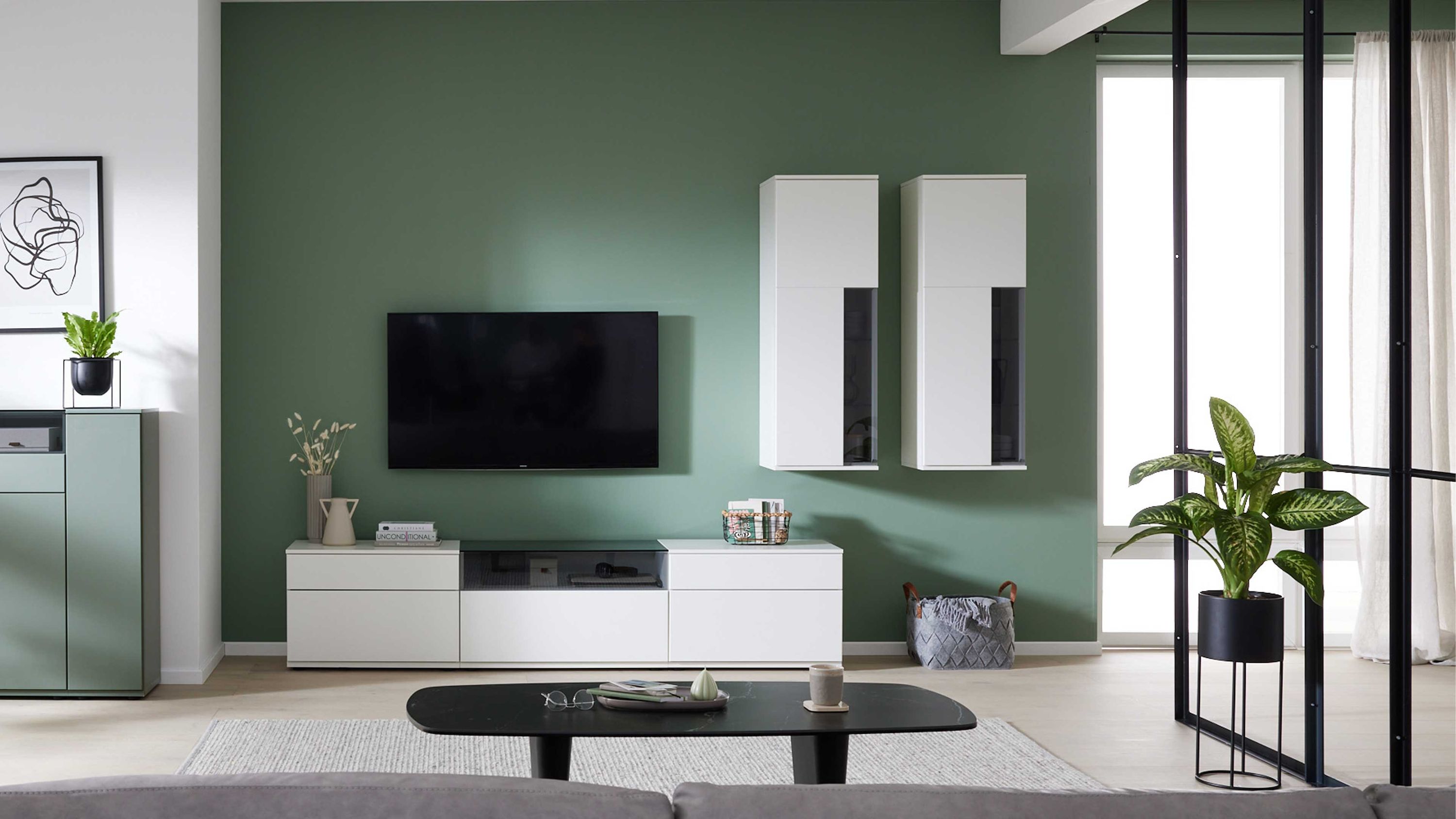 Weiße Wohnwand mit grauen Glastüren und Fernseher Wohnwand mit weißen Schrankelementen und grauen Glastüren an grüner Wand mit Fernseher