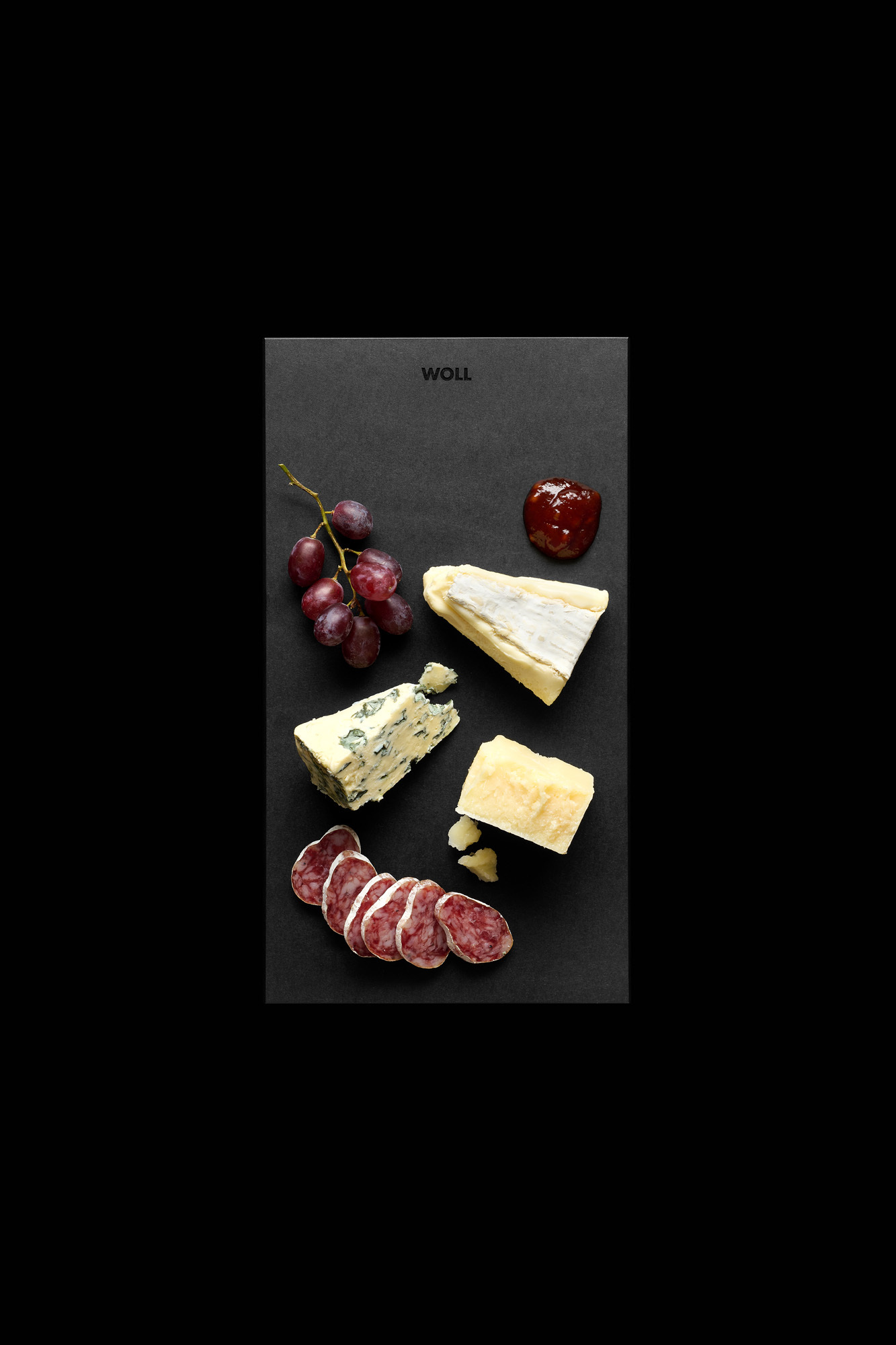 Schwarzes Servierbrett mit Trauben, verschiedenen Käsesorten, Salami und rotem Chutney vor schwarzem Hintergrund angerichtet