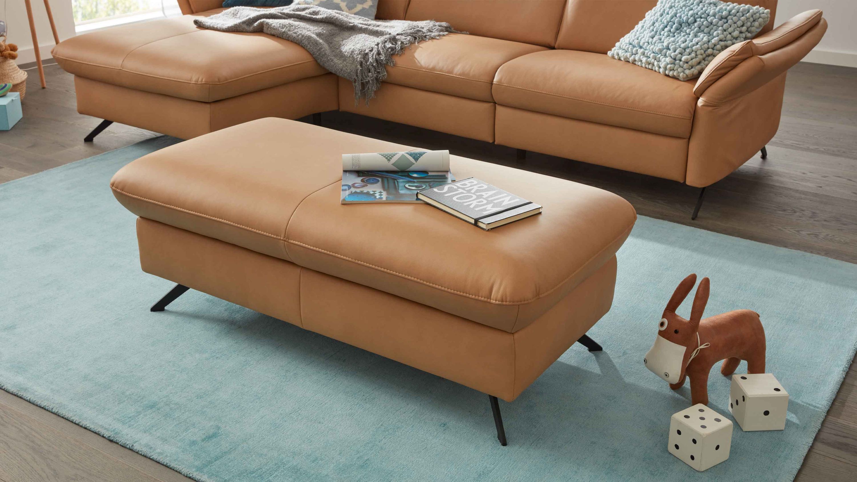Großer rechteckiger Hocker in honigfarbenem Leder mit schwarzen Metallfüßen auf blauem Teppich vor passendem Sofa