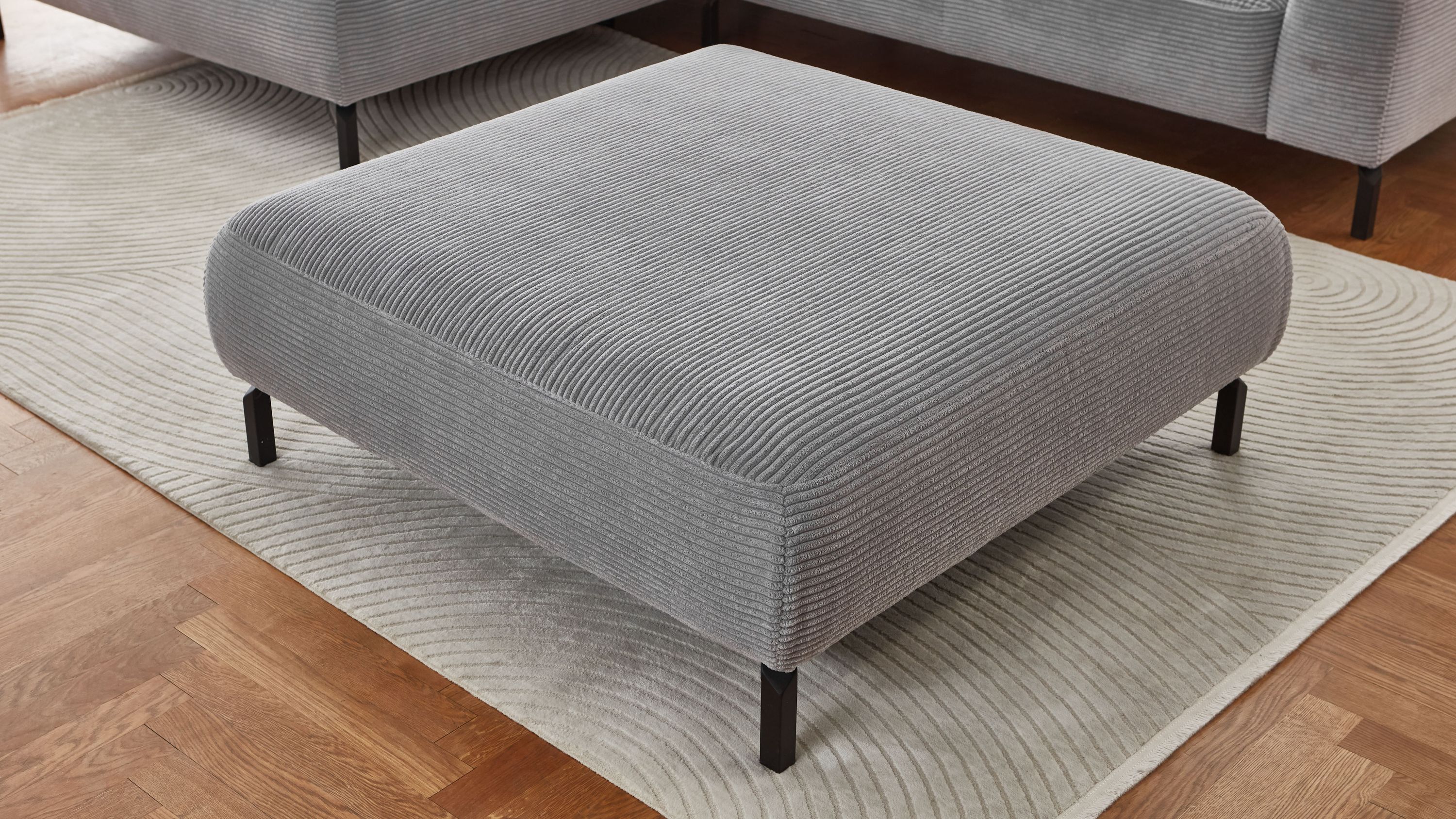 Quadratischer Hocker mit grauem Cordbezug und schwarzen Beinen auf hellem Teppich