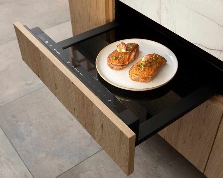 Wärmeschublade mit Fleischstückchen in Bora Küche Zwei Fleischstücke in einer geöffneten Wärmeschublade mit Holzfront, moderner Küchenboden