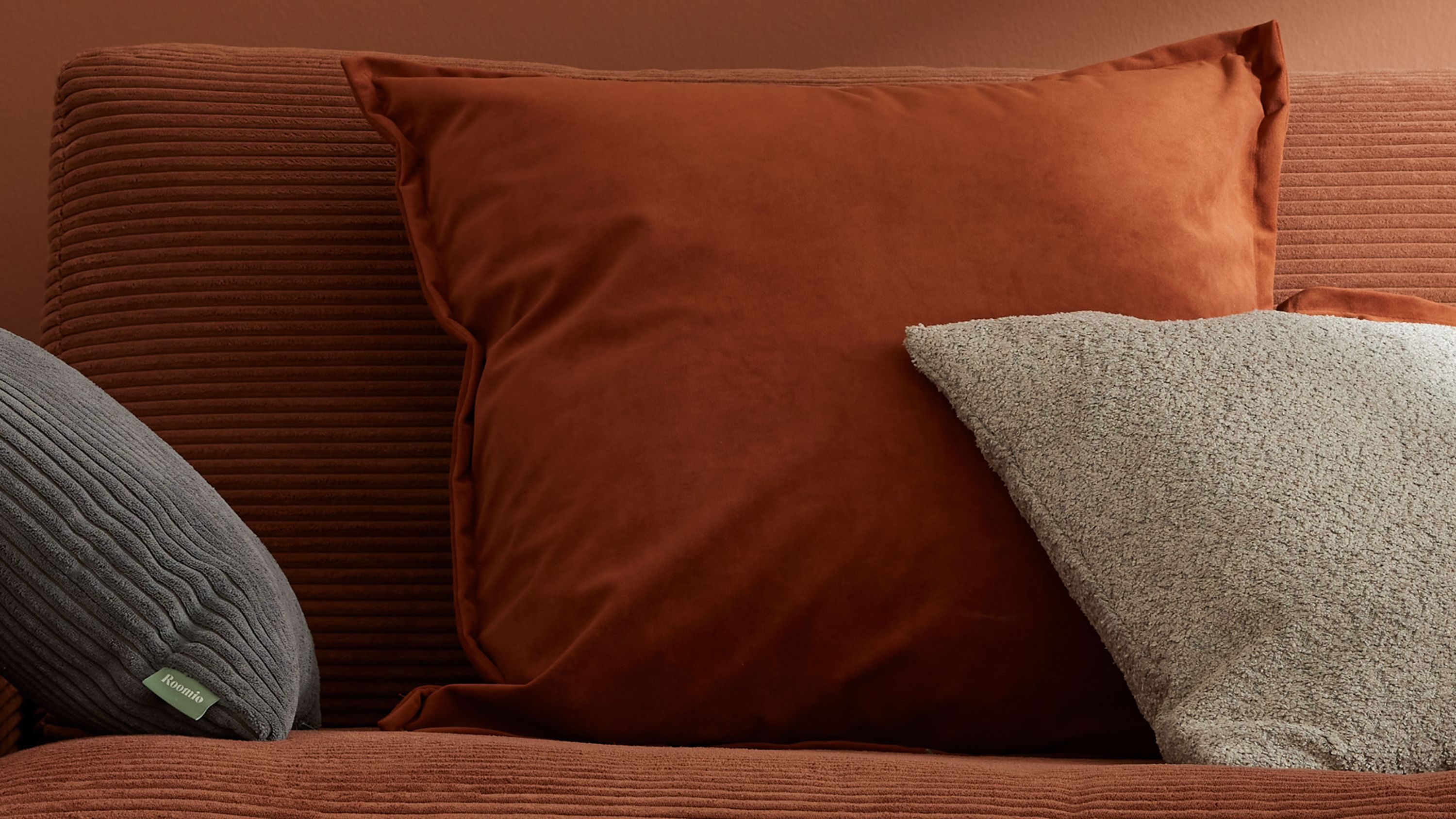 Quadratisches rostfarbenes Kissen auf Sofa neben grauem Kissen mit strukturierter Oberfläche.