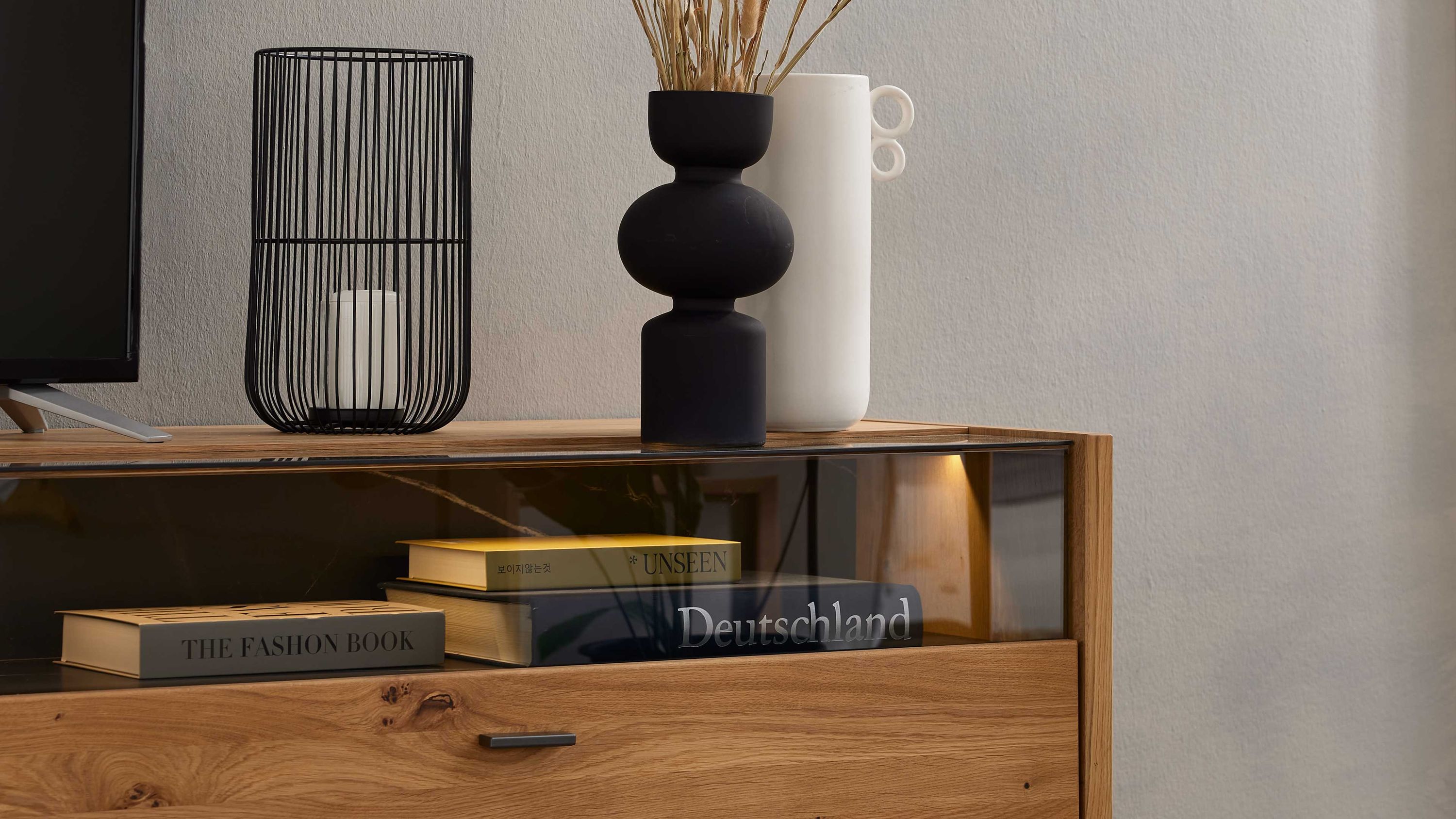 Wohnzimmerschrank mit Holzfront Glastür und integrierter Beleuchtung darauf zwei Vasen und ein Kerzenhalter