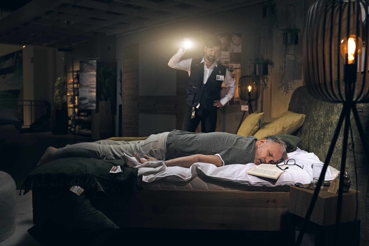 Mann schläft auf Bett im Möbelhaus Mann liegt schlafend auf einem Bett im Möbelhaus, ein Mitarbeiter inspiziert mit Taschenlampe.