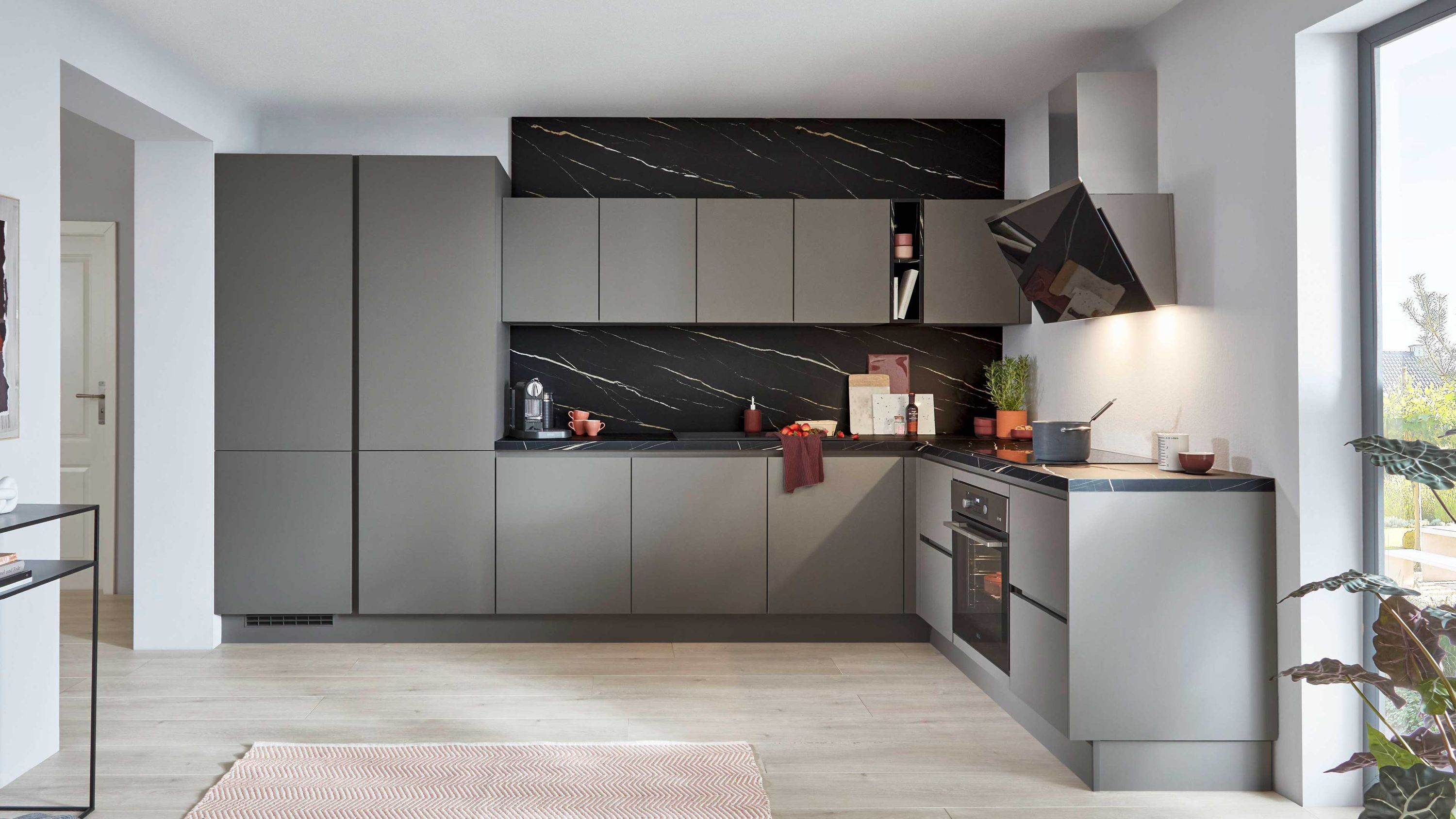 Graue L-Küche mit Marmor-Rückwand und Einbaugeräten. Graue L-Küche mit grifflosen Fronten schwarzer Marmor-Rückwand Einbaugeräten und moderner Dunstabzugshaube.