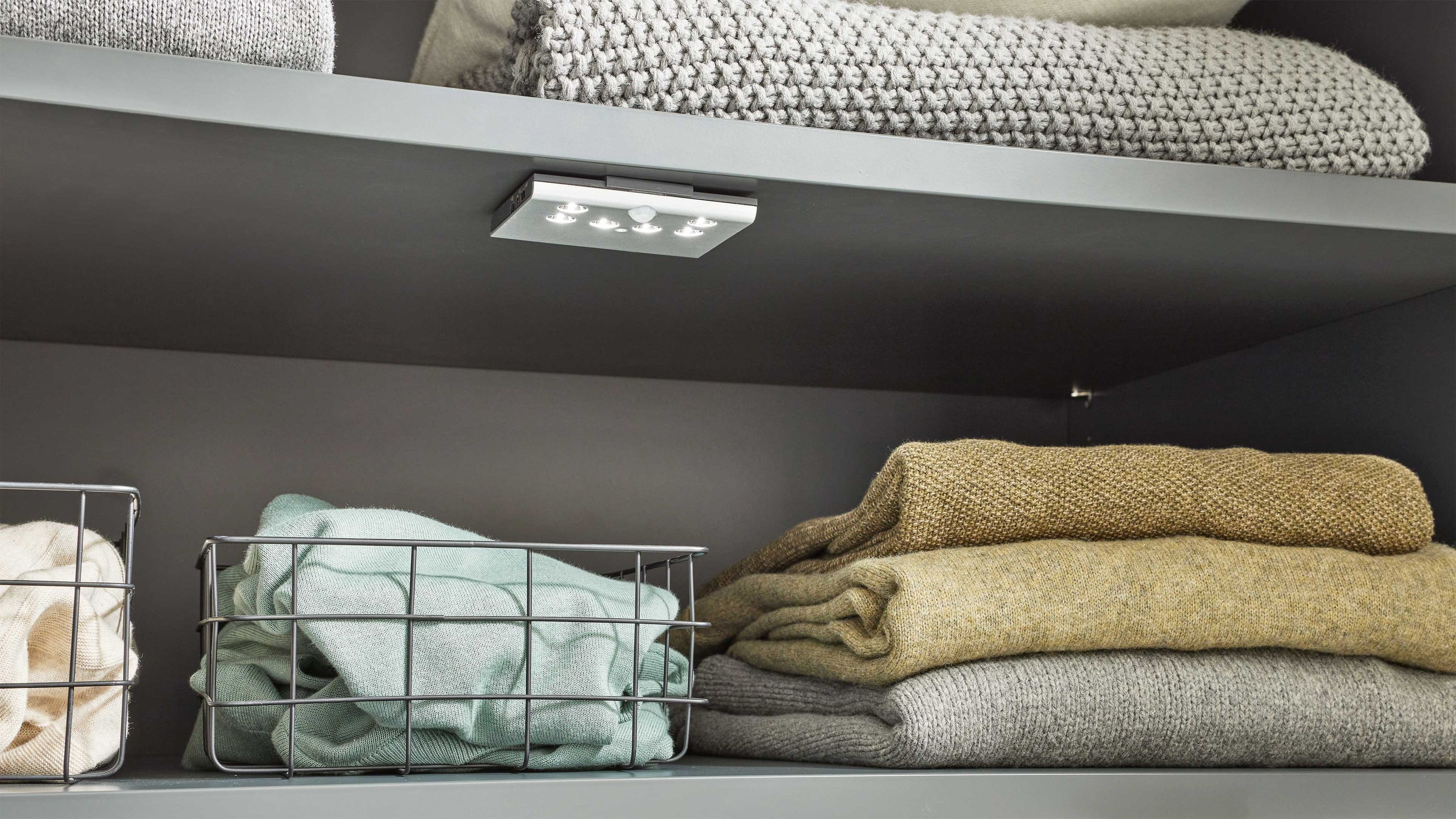 Weiße LED-Leuchte unter Regal mit Decken und Körben Rechteckige weiße LED-Leuchte unter einem Regal mit gefalteten Decken und Drahtkörben