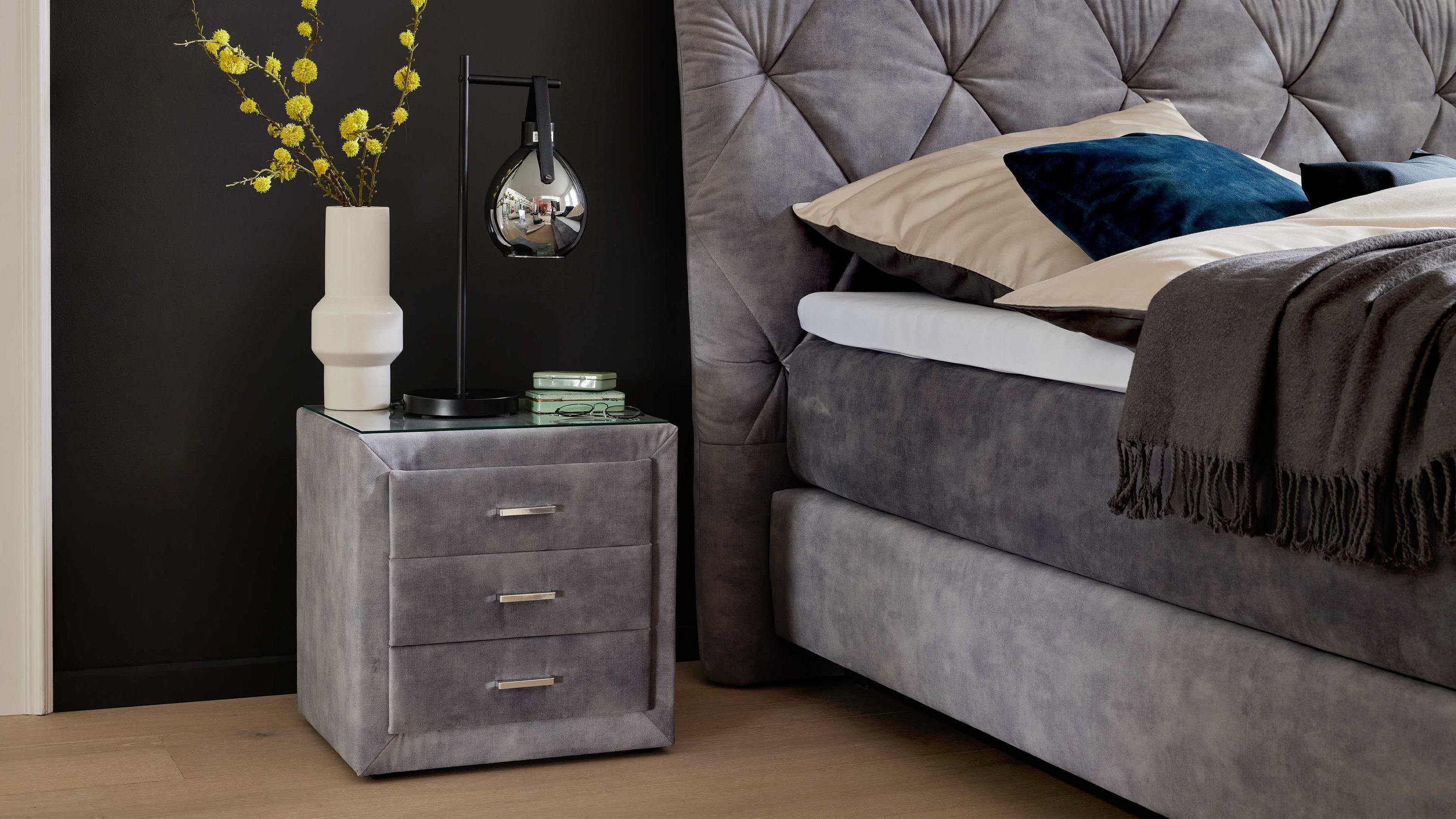 Silberfarbene Nachtkonsole mit drei Schubladen und Glasplatte neben grauem Boxspringbett