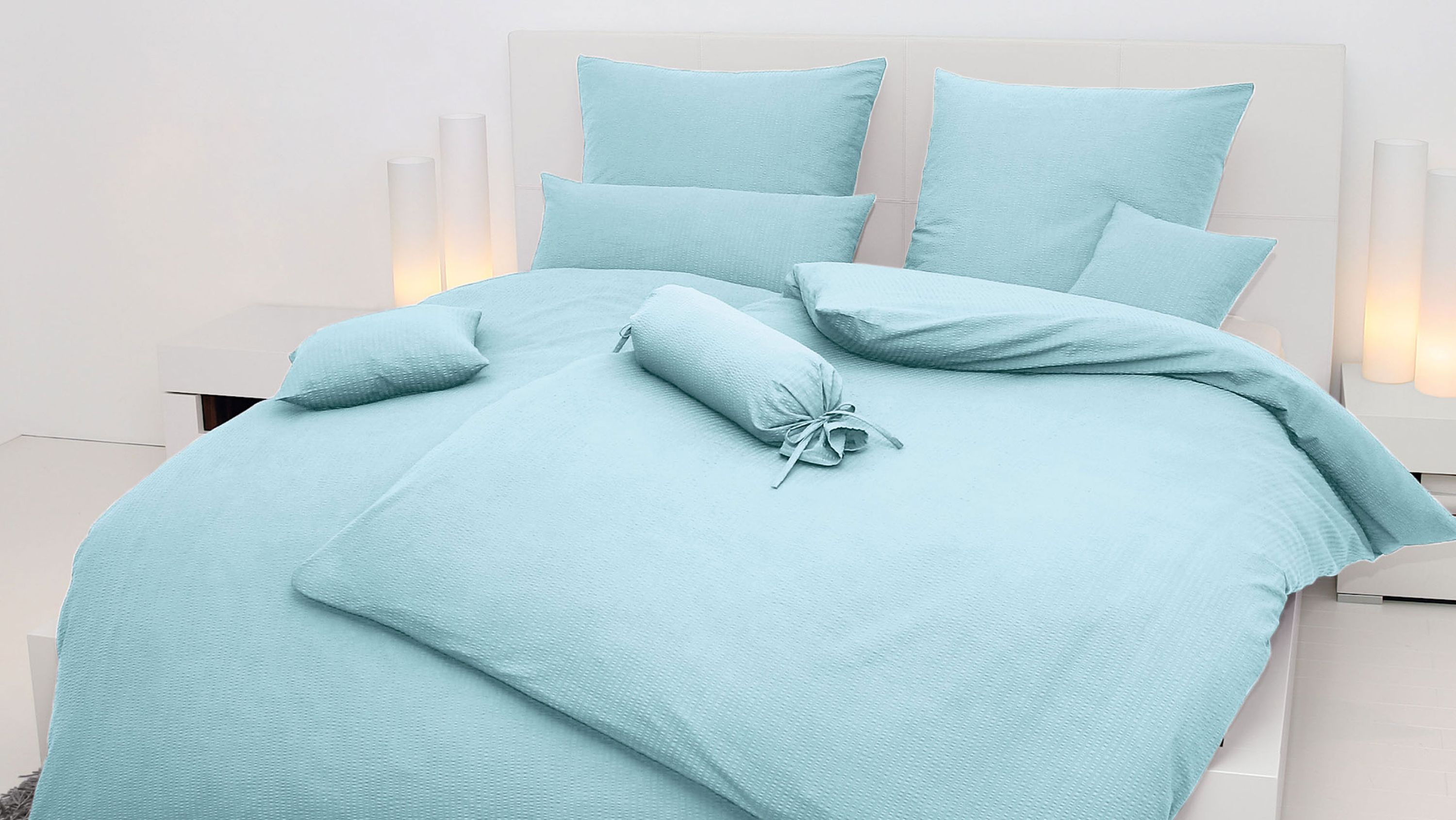 Hellblaue Bettwäsche mit mehreren Kissen und einem runden Kissen auf einem weißen Bett