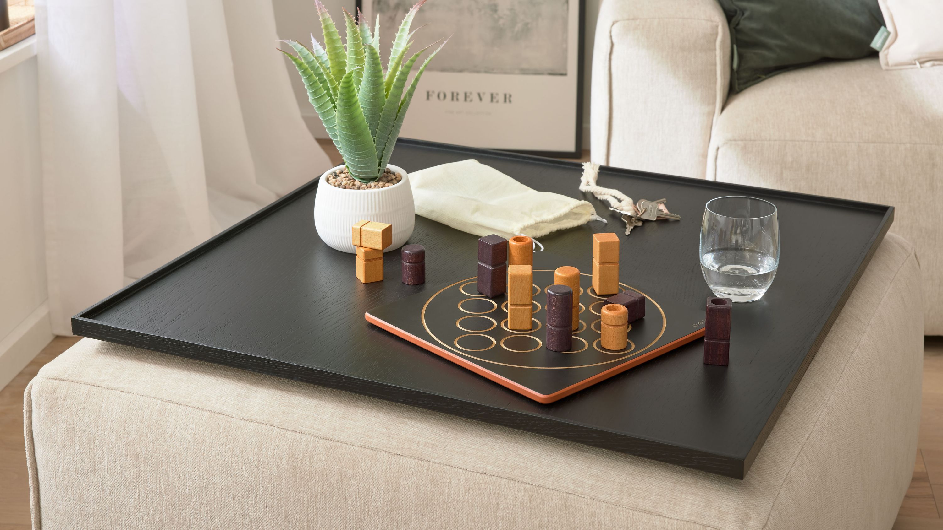 Schwarzes Tablett mit Spielbrett Glas Schlüsselbund und Blumentopf auf hellem Hocker neben rotem Sofa