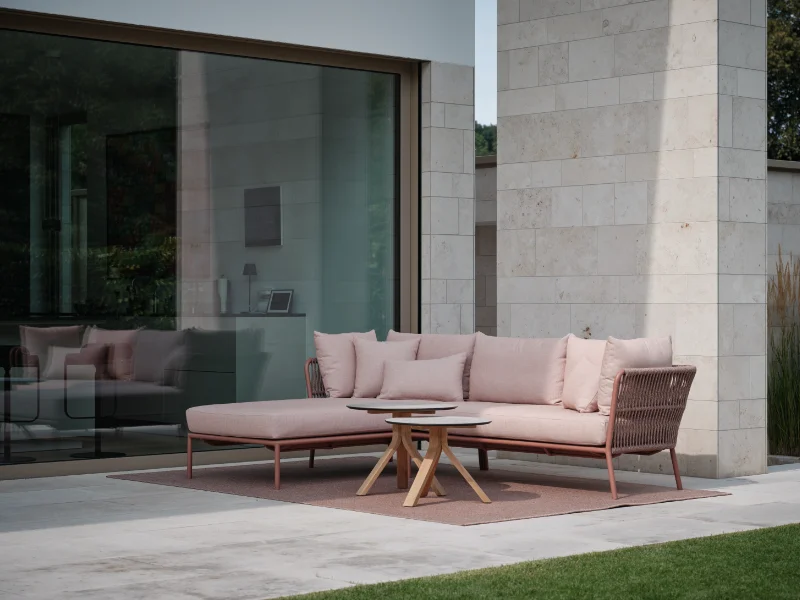 Moderne Outdoor-Lounge mit L-förmigem Sofa in Rosa und rundem Couchtisch auf Terrasse vor Steinfassade.