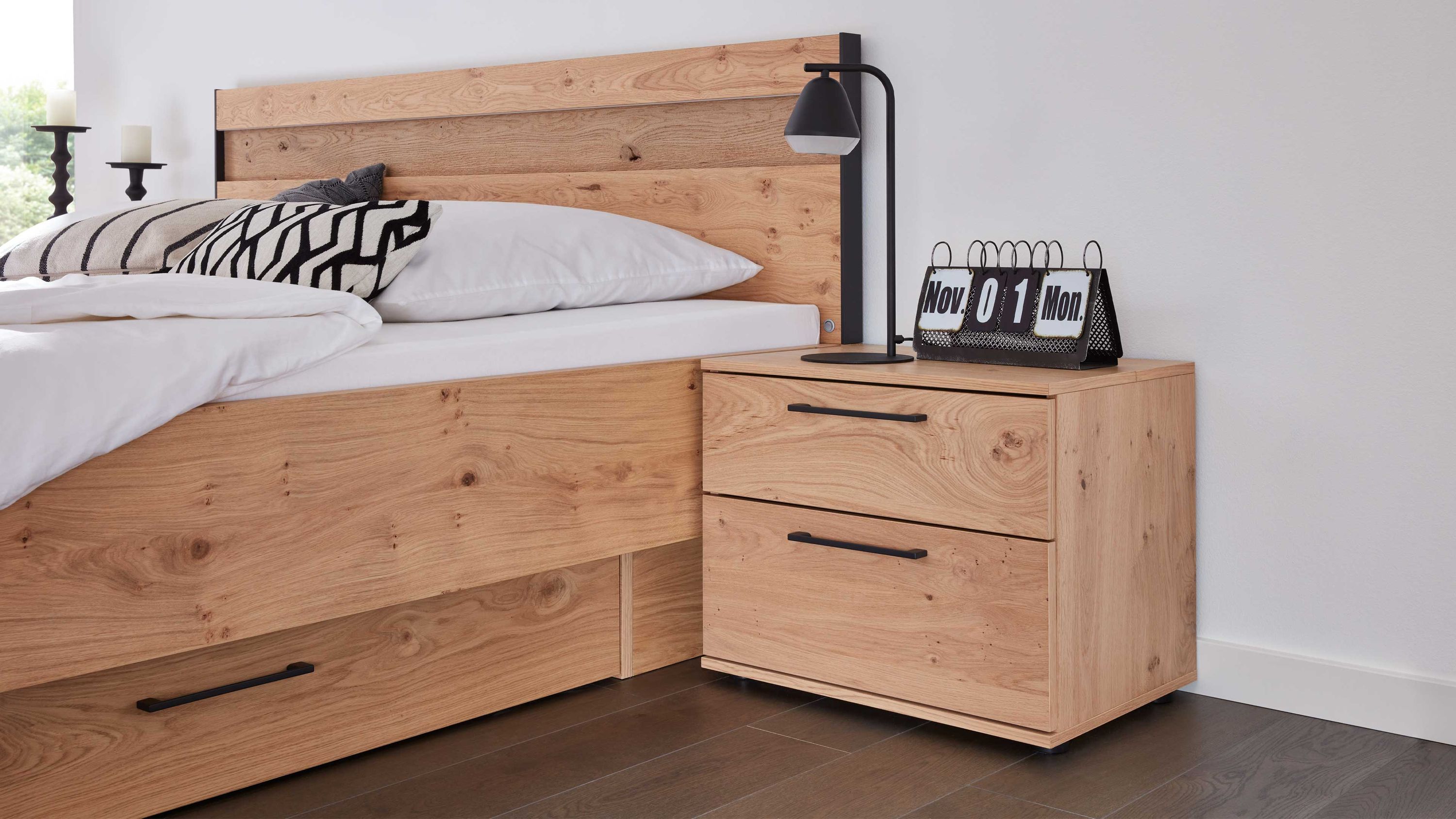 Nachtkommode mit zwei Schubladen in hellbrauner Holzoptik neben einem Bett mit Kopfteil aus Holz