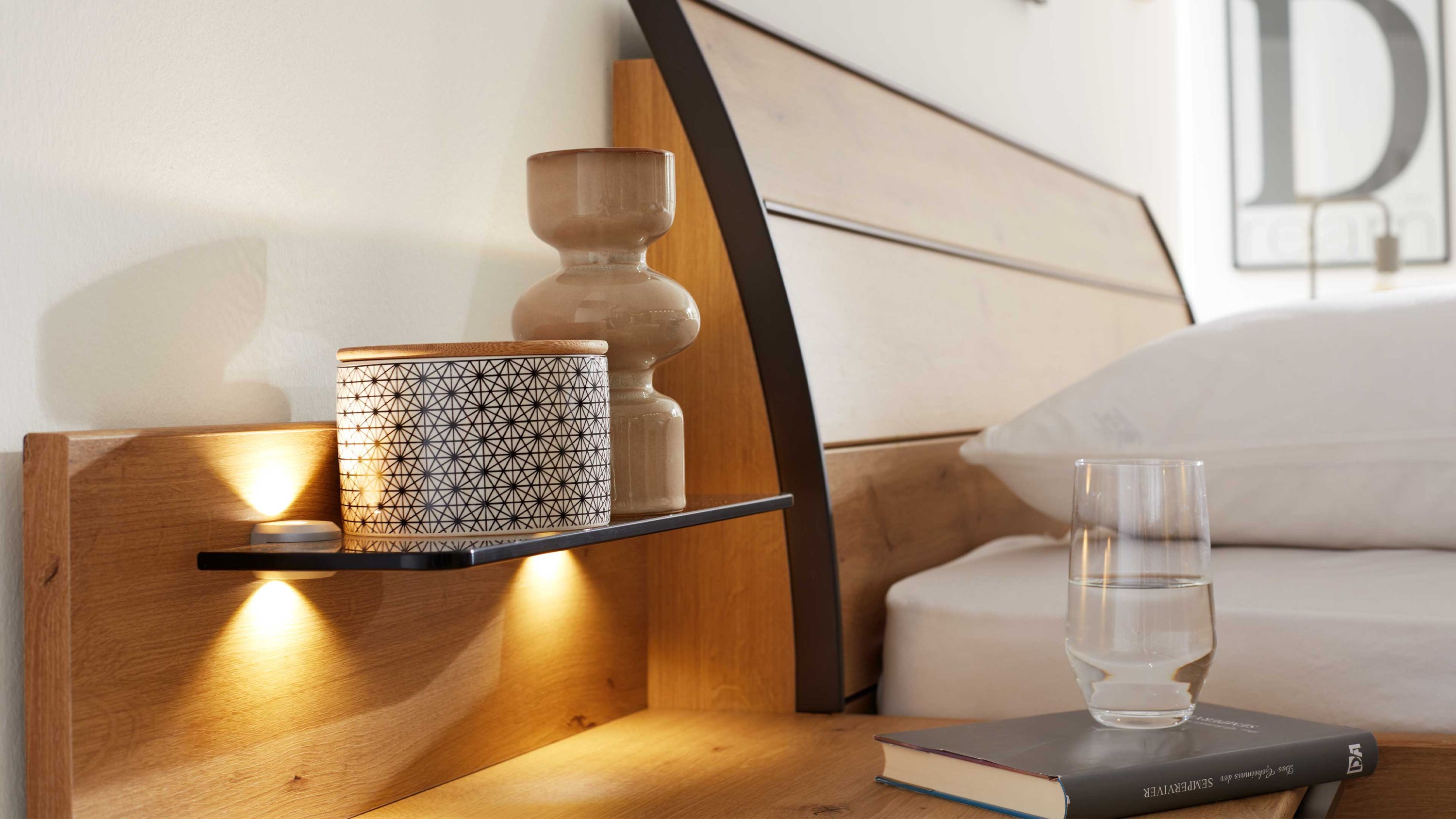 Paneel-Set mit Regal und Beleuchtung aus Holz Zweiteiliges Paneel-Set aus Holz mit schwarzem Regalbrett und integrierter Beleuchtung neben einem Bett