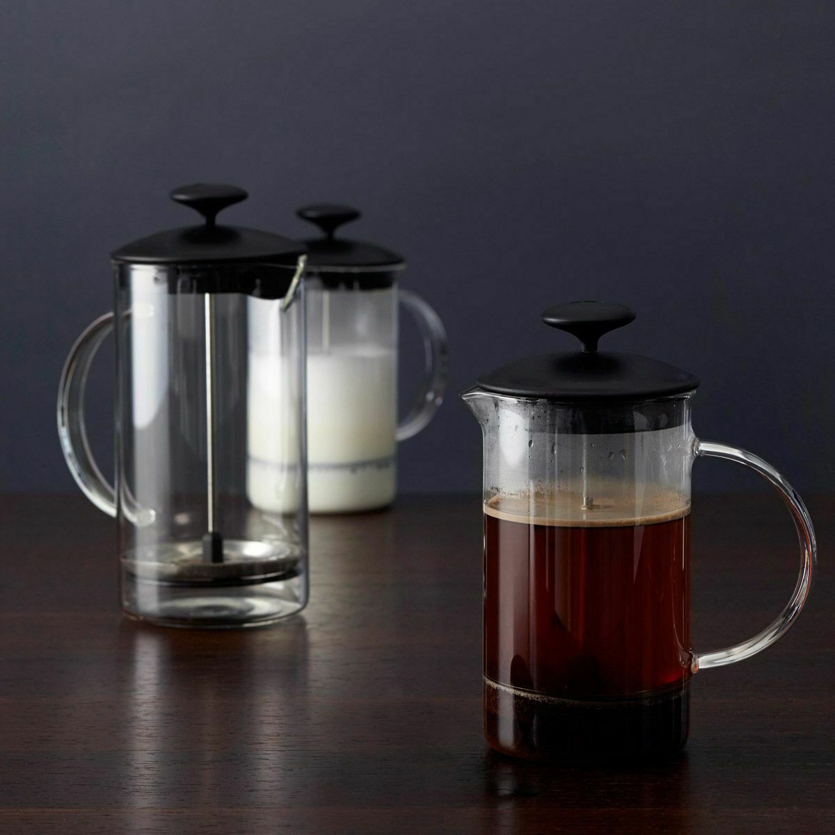 Drei French Press Kannen auf dunkler Fläche – eine mit Kaffee, eine mit Milch, eine leer, frontal fotografiert