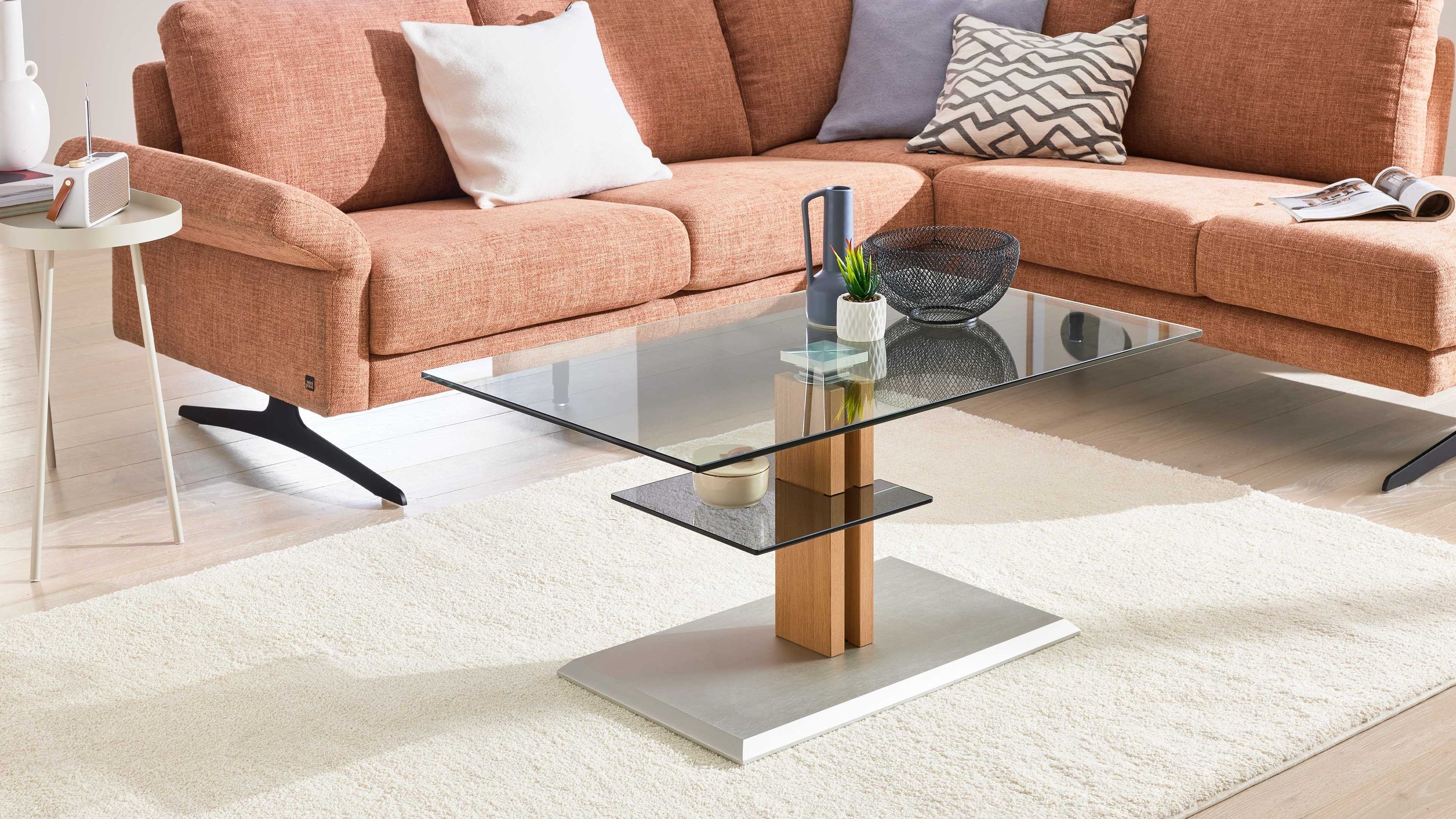 Rechteckiger Couchtisch mit klarem Glas und Holzsäule auf rechteckiger Metallplatte vor braunem Ecksofa