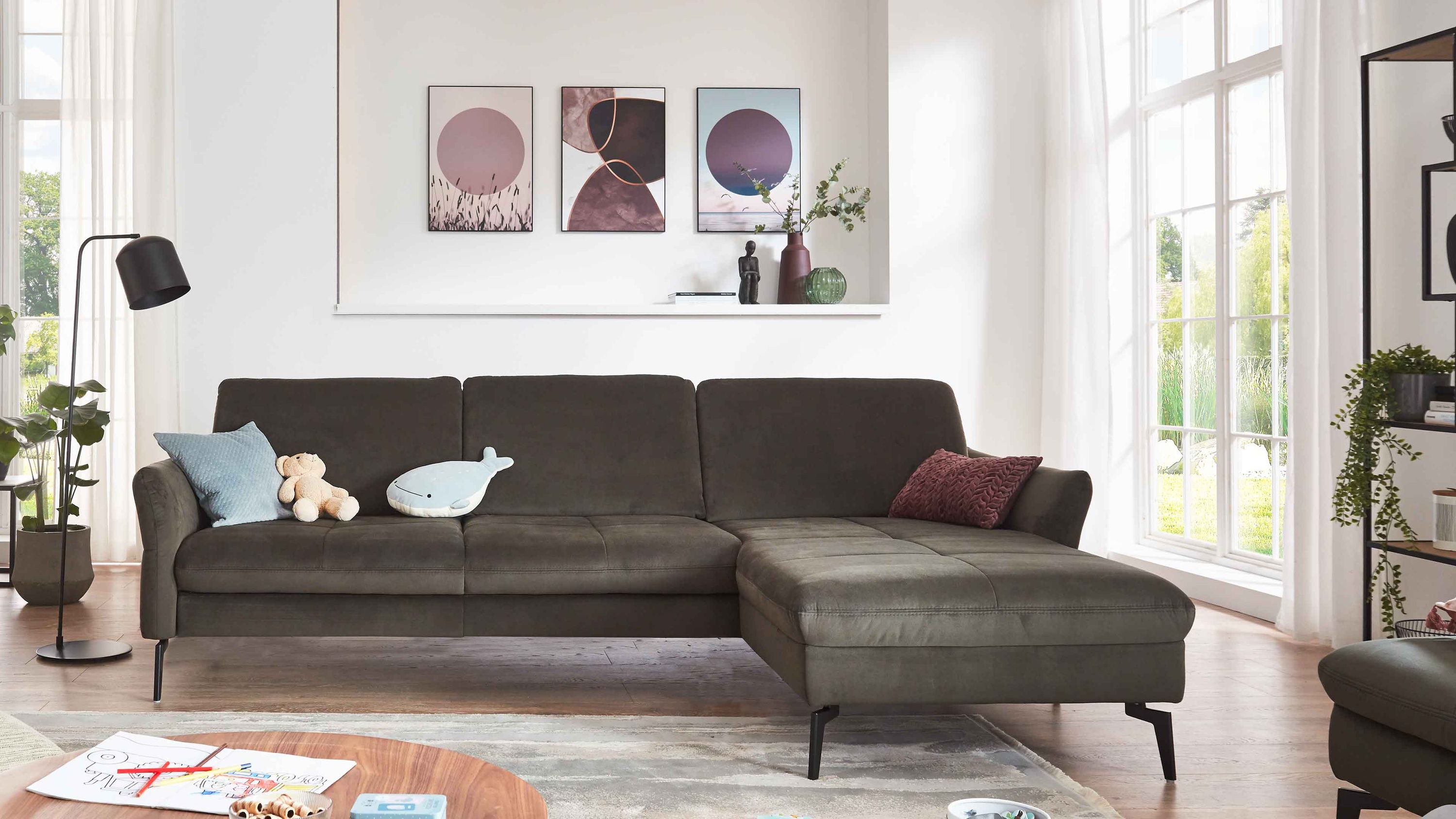 Ecksofa mit olivgrünem Stoffbezug und schwarzen Metallfüßen in hellem Wohnzimmer mit Dekokissen und Kuscheltieren