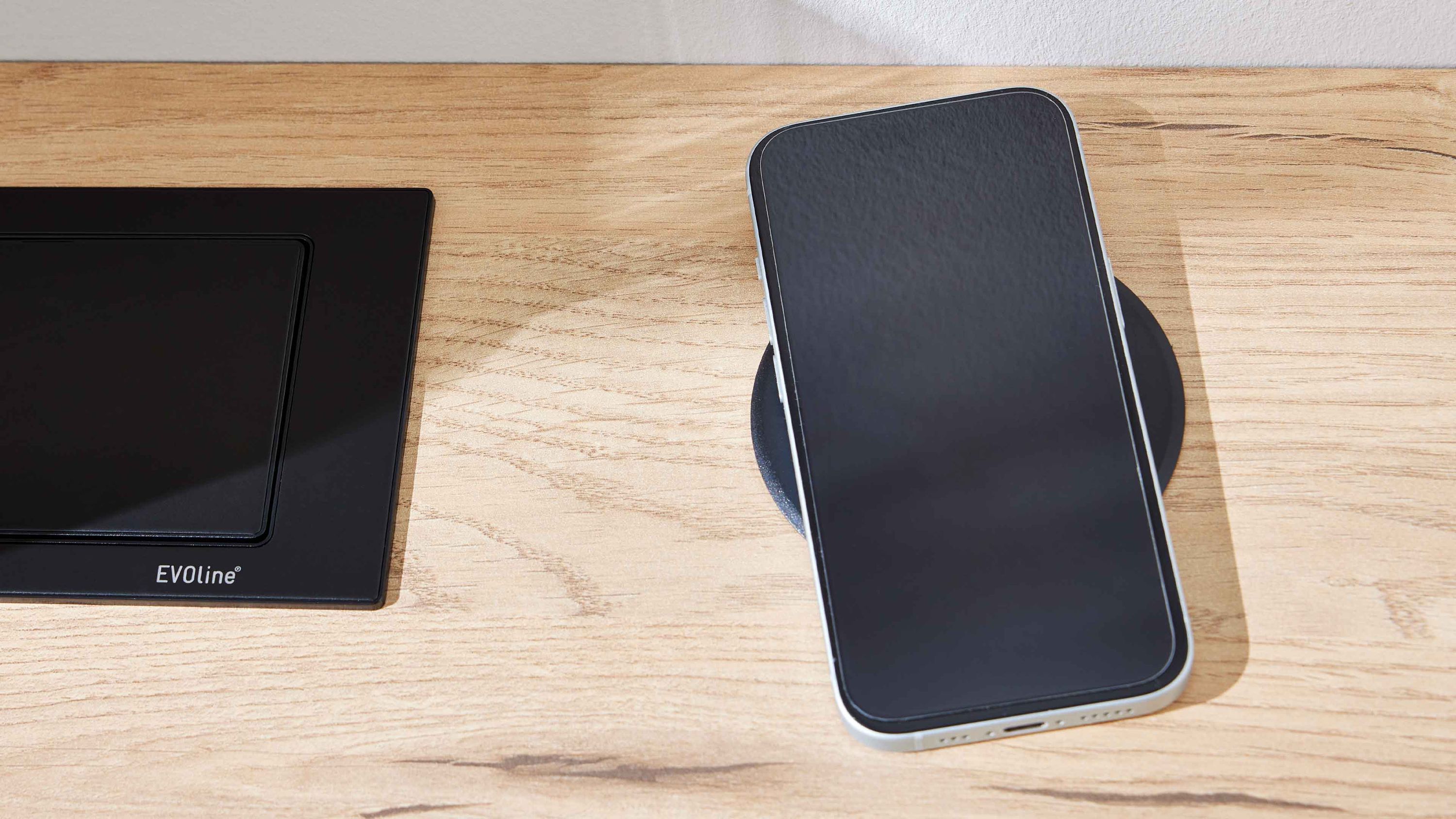 Schwarzes Smartphone liegt auf kabellosem Ladegerät auf hellem Holzschreibtisch