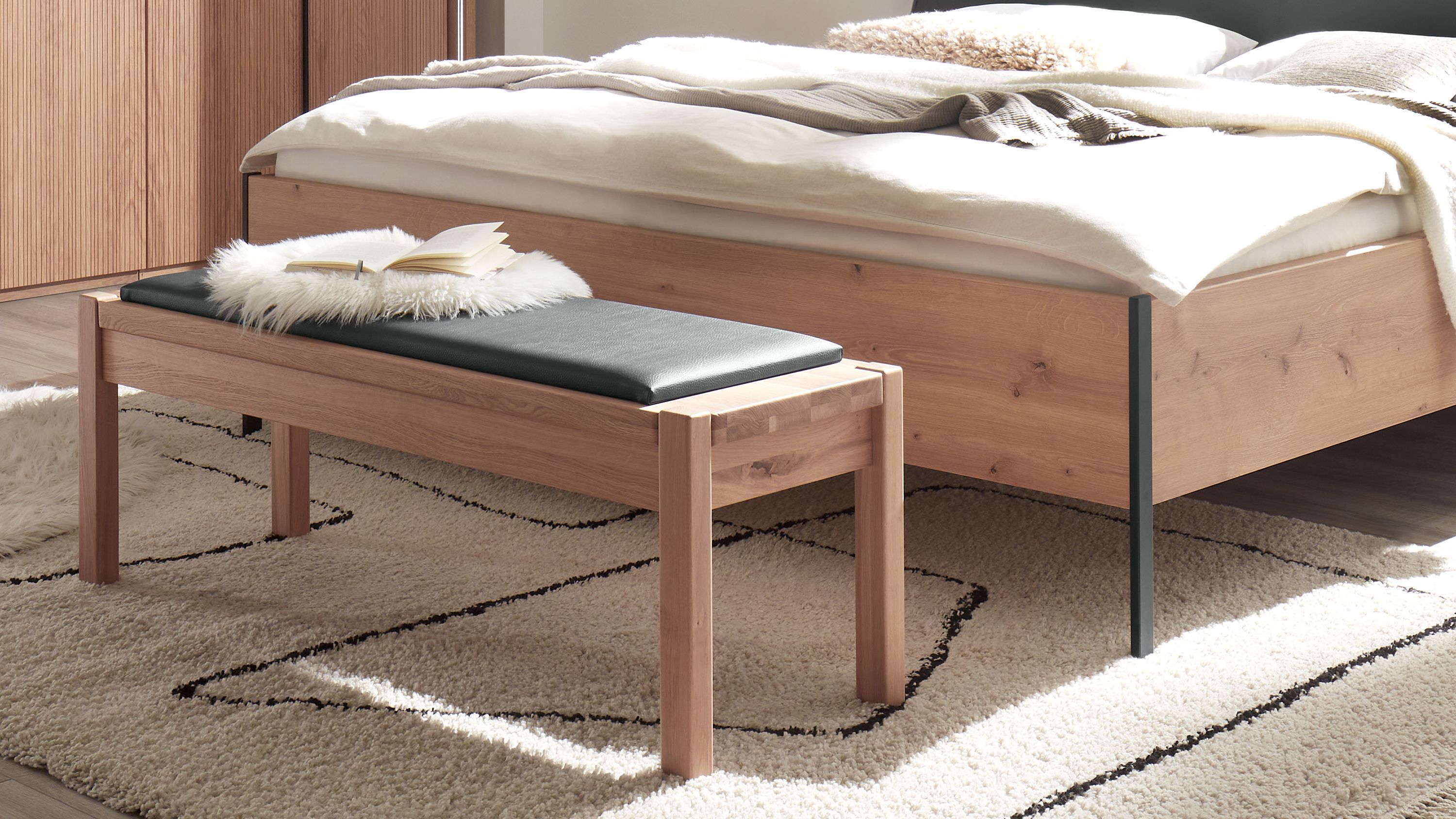 Bank mit gepolsterter Sitzfläche in dunkler Farbe vor einem Bett aus hellem Holz in einem Schlafzimmer