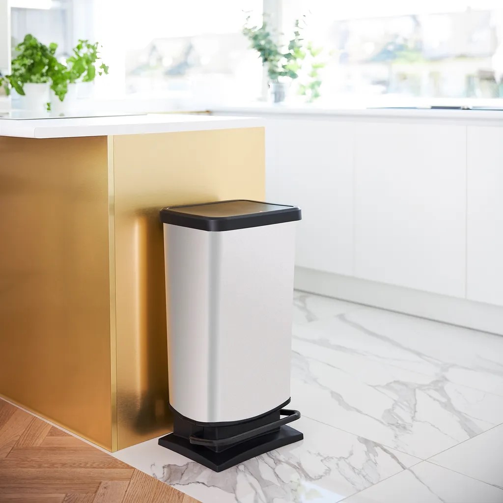 Silberfarbener Treteimer mit schwarzem Deckel in moderner Küche Treteimer silberfarben mit schwarzem Deckel in moderner Küche neben goldenem Schrank schräg erhöht fotografiert