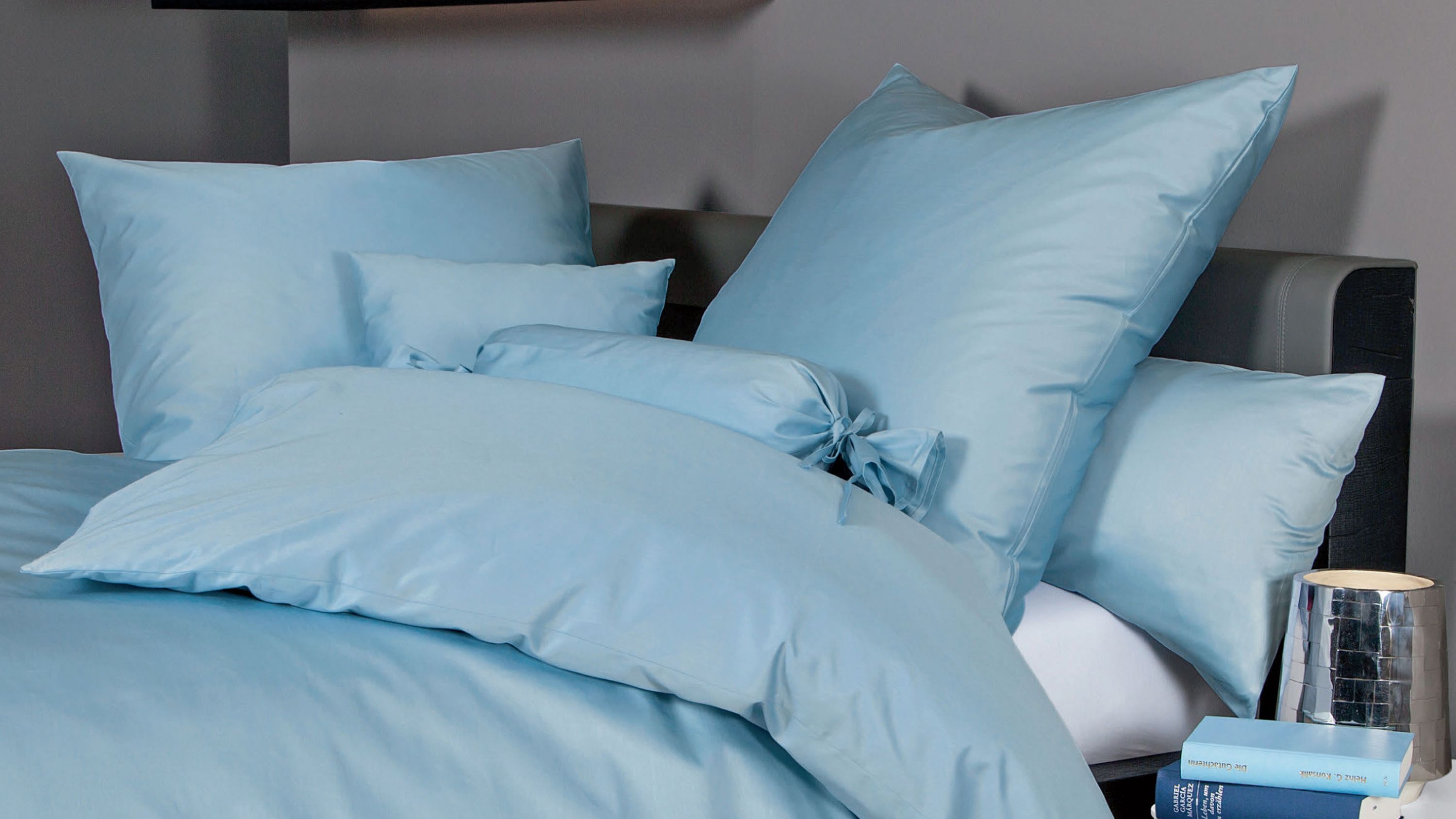 Hellblaue Bettwäsche mit mehreren Kissen auf einem Bett mit grauem Kopfteil