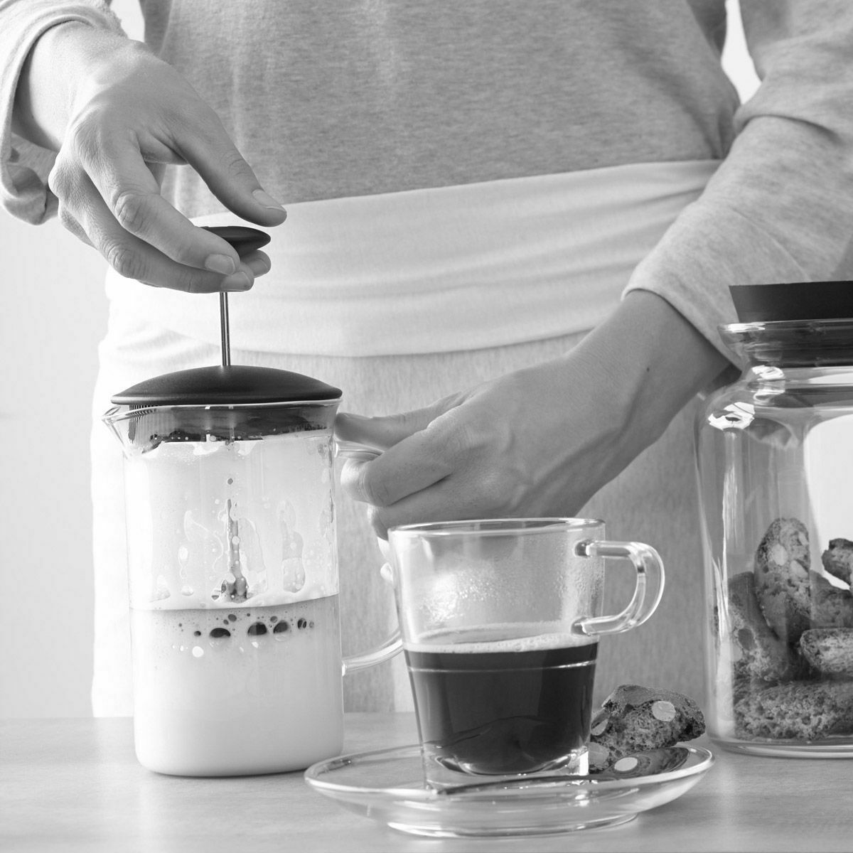 Person schäumt Milch mit Handaufschäumer neben Kaffeetasse und Keksdose auf, aus Sicht leicht schräg von oben.