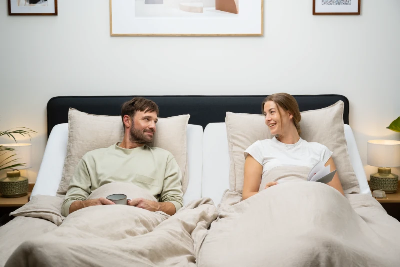 elles, modernes Schlafzimmer mit komfortablem Boxspringbett, zwei entspannt liegenden Personen unter heller Bettwäsche, abgestimmten Kissen, Nachttischen mit Beleuchtung und ruhiger Wohnatmosphäre.