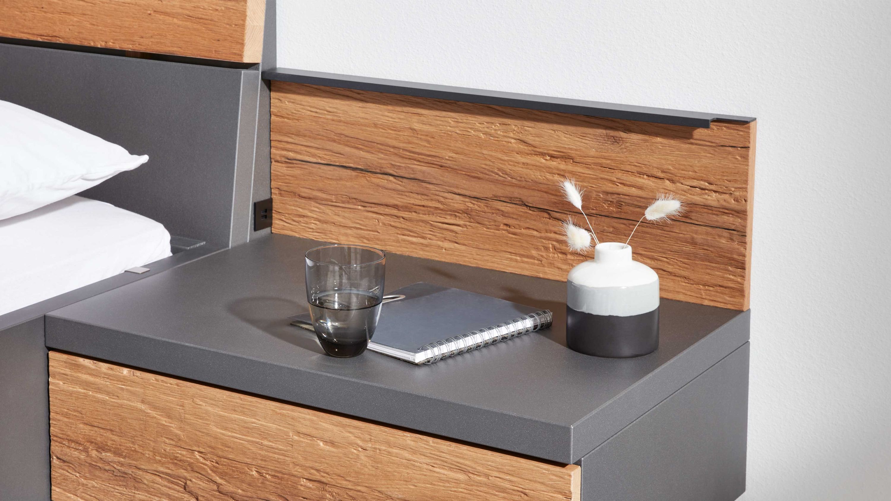 Aufsatzpaneel mit Holzoptik in braun und grauer Ablagefläche neben Bett mit Glas Notizbuch und Vase