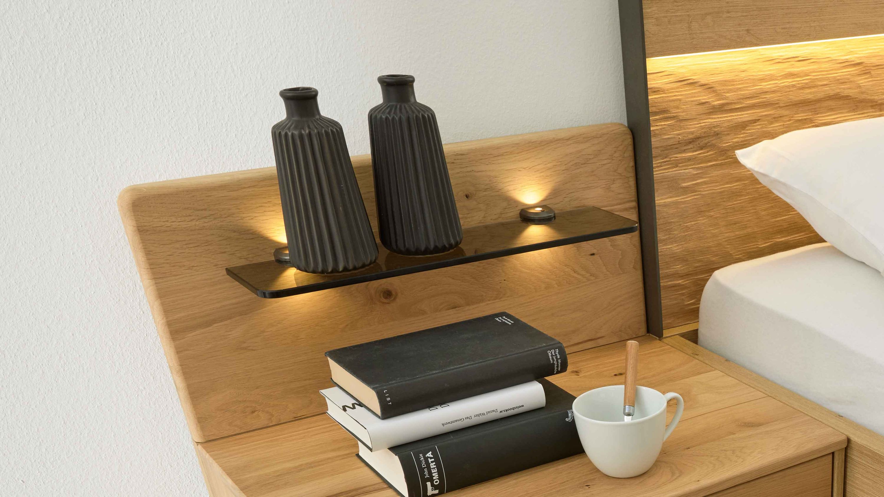 Paneel-Set mit Holzrückwand und Glasablage Zweiteiliges Paneel-Set mit Holzrückwand schwarzem Glasregal und integrierter Beleuchtung neben einem Bett