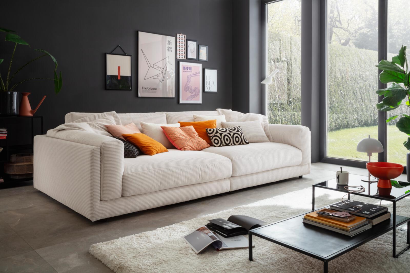M4840142953729601_000 Großes, gemütliches Sofa von Candy Polstermöbel in hellem Stoff mit bunten Kissen, vor dunkler Wand mit moderner Bildergalerie - Mit Link zur Markenseite Candy Polstermöbel