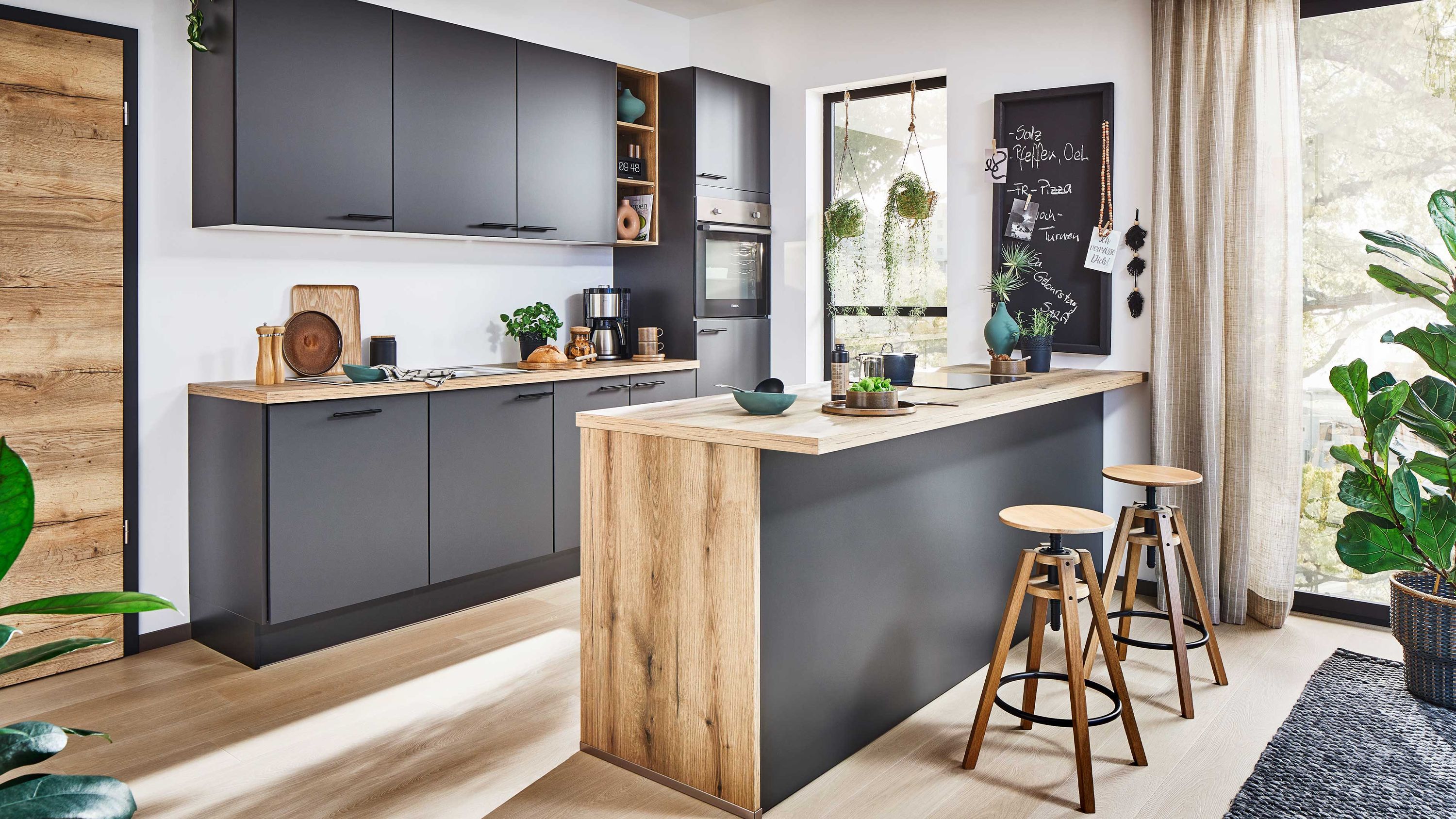 Dunkelgraue Küche mit Holzinsel und Kreidetafel Dunkelgraue Küche mit Holzarbeitsplatte Insel mit Hockern Pflanzen Fenster Wandtafel Tageslicht.