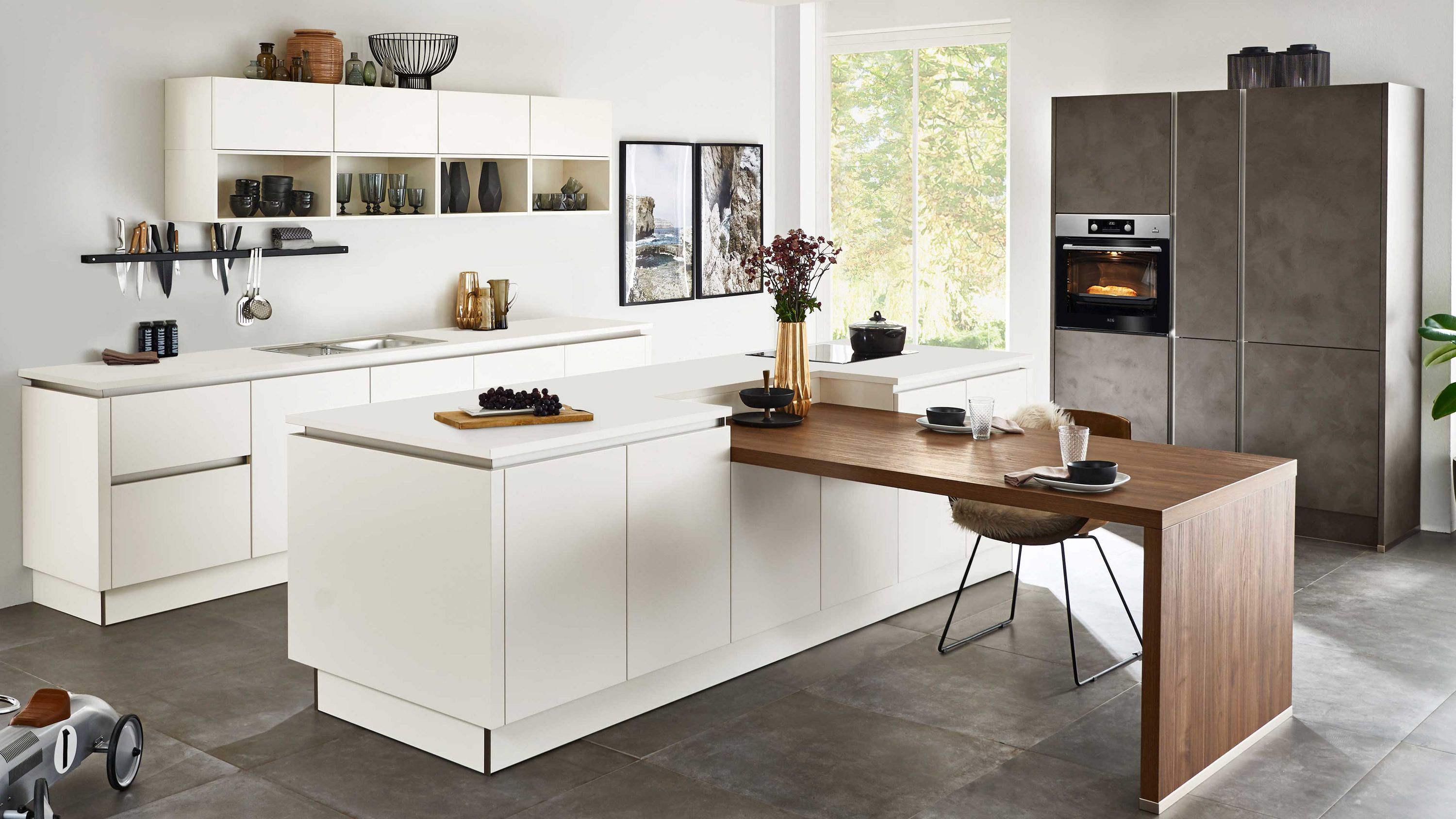Moderne Küche mit weißer Insel und integriertem Holztisch Küche mit weißen Schränken Holz-Esstisch an Insel Einbauofen Regalen Tageslicht durch großes Fenster