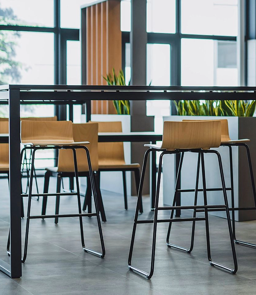 Hochtisch mit Barstühlen in moderner Büro-Pausenzone, große Fenster, Pflanzen und offene Gestaltung im Office-Design