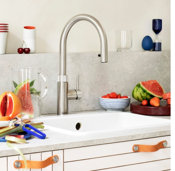 Moderne Küchenarmatur aus Edelstahl mit Auslauf über weißem Spülbecken, umgeben von Obst, Gläsern und farbenfrohem Zubehör.